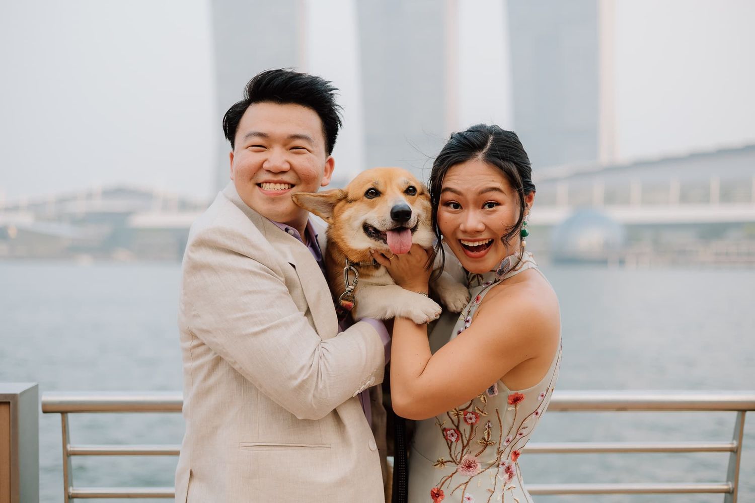 A happy couple poses with their smiling Shiba Inu dog against a waterfront backdrop.