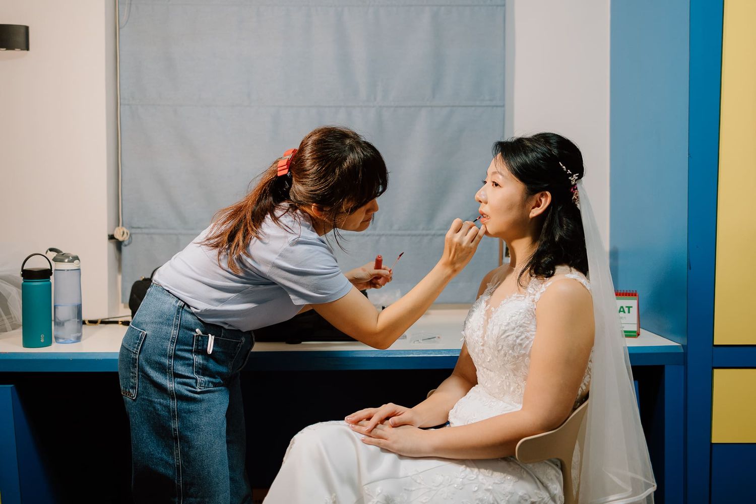 Professional stylist applies makeup to client while seated in bright blue and white salon setting.