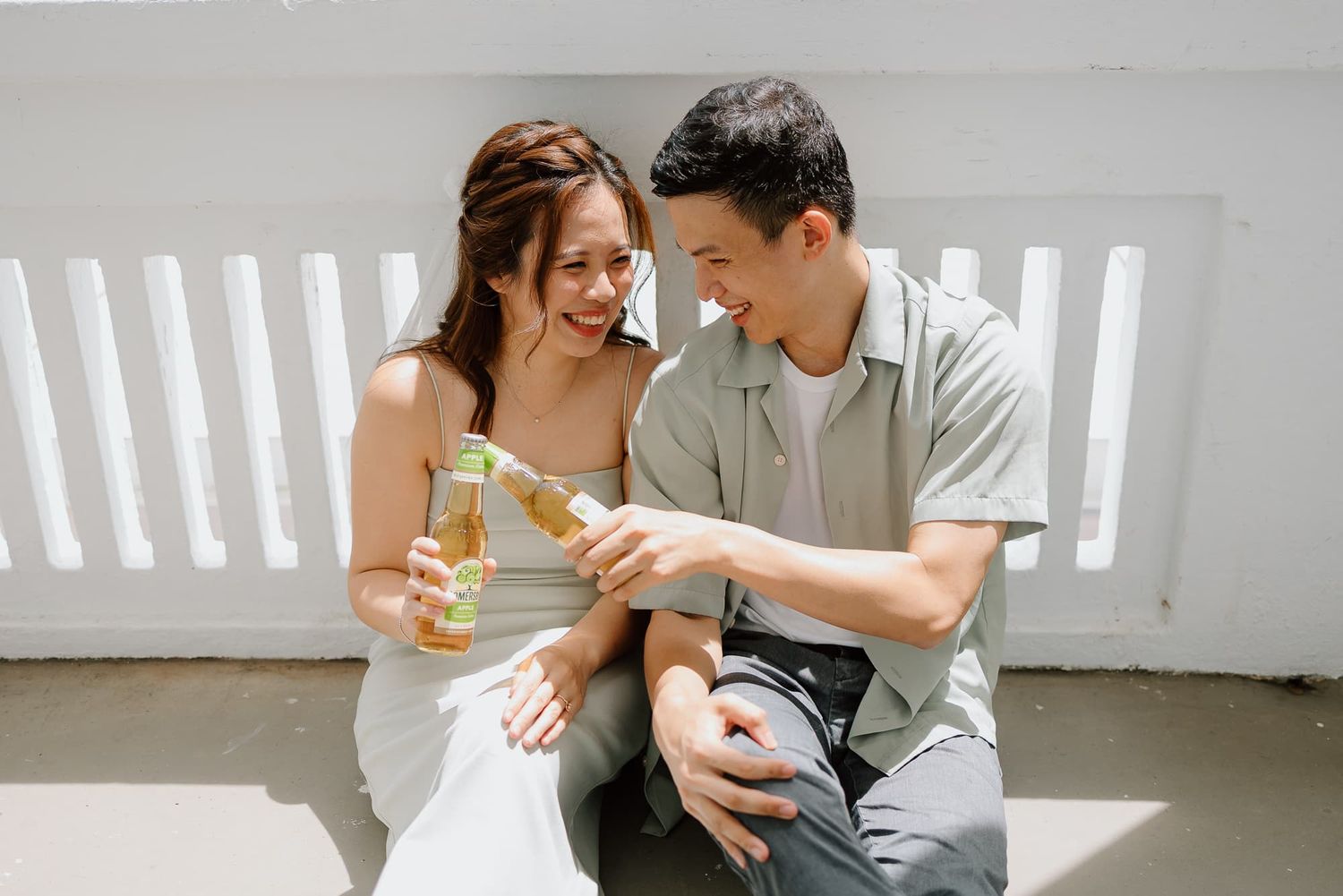 Couple sharing drinks and laughter while sitting on a white bench in bright sunlight.