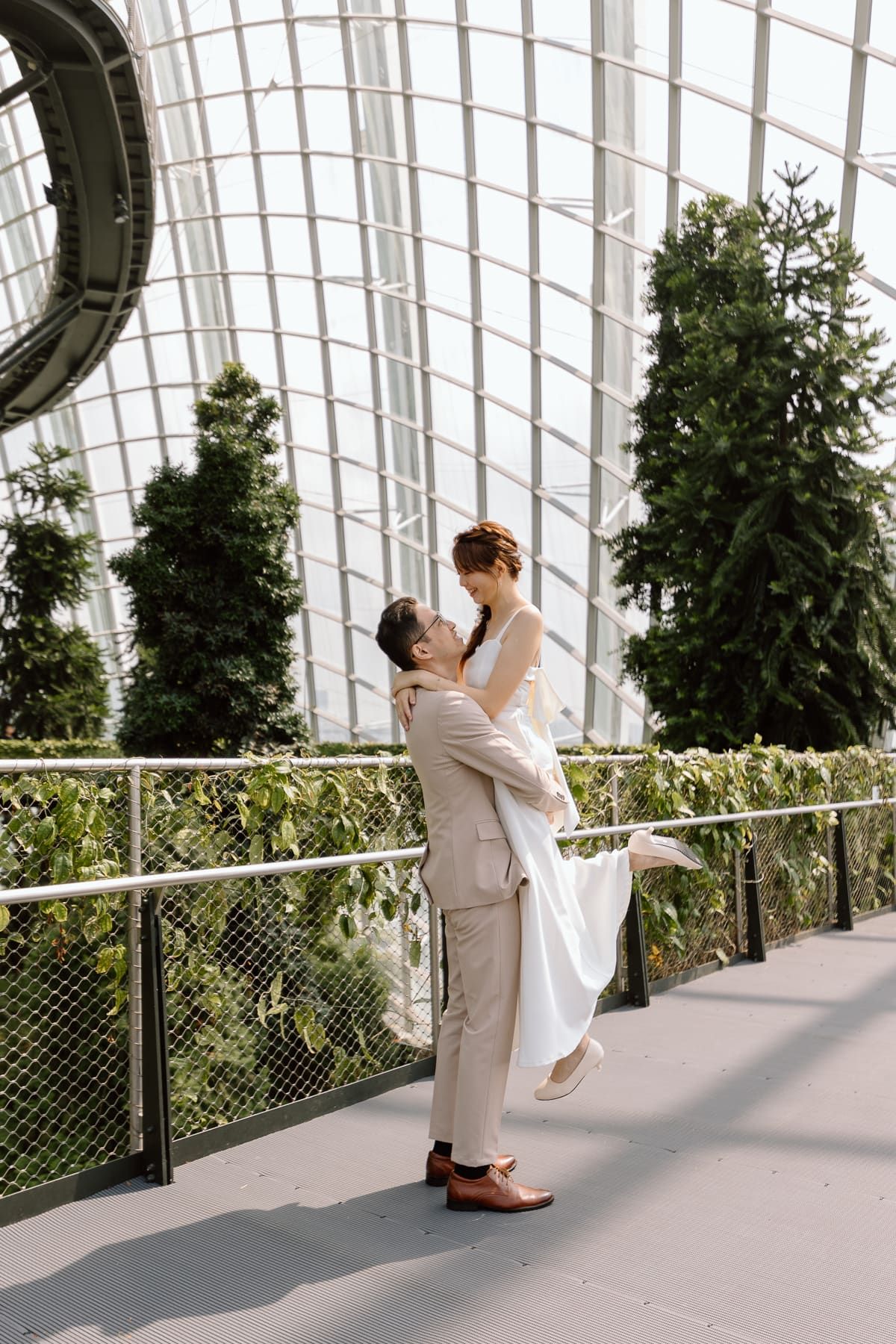 A romantic lift pose captured in a glass-domed conservatory with evergreen trees.