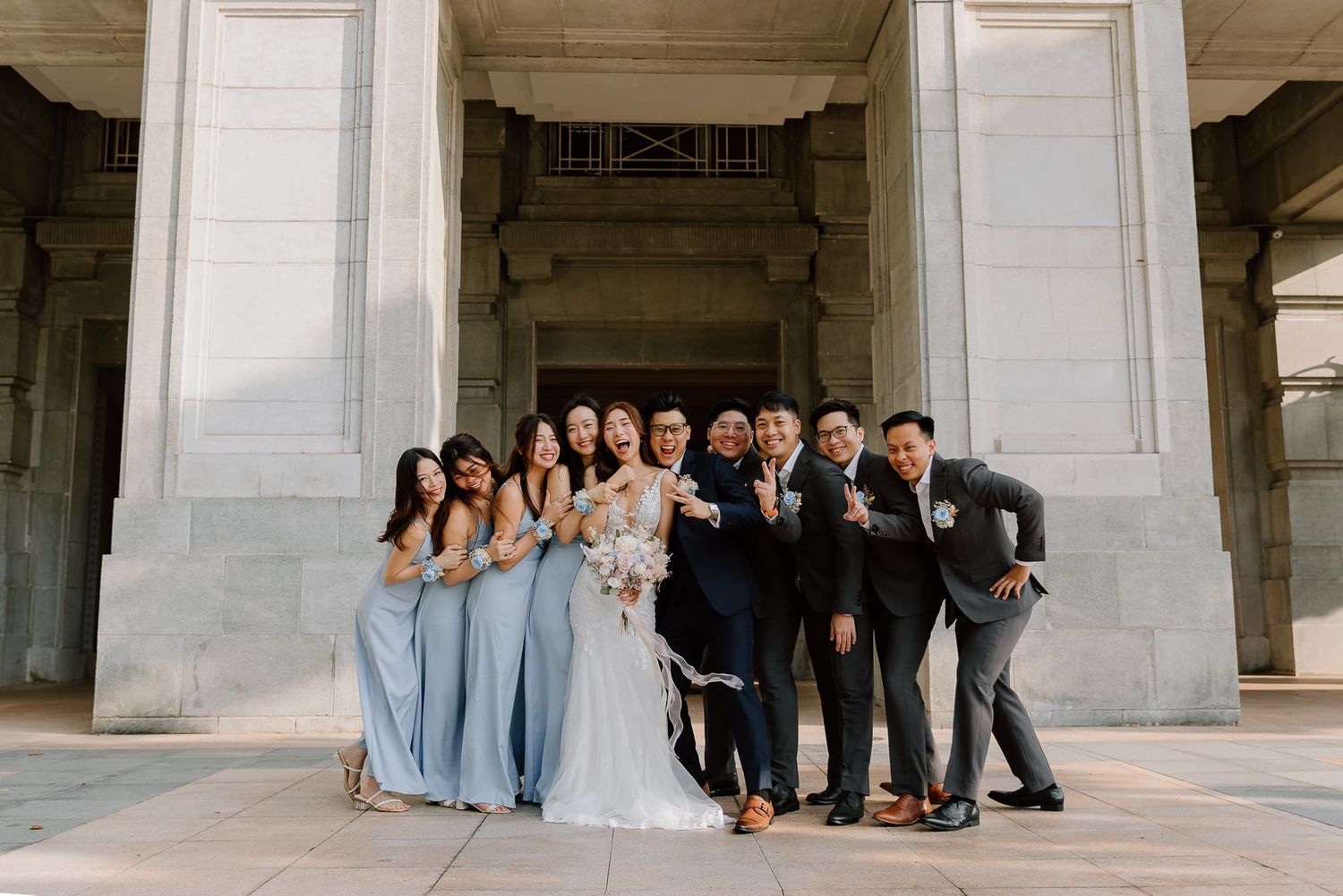 Wedding party in light blue dresses and dark suits posing together under a classical stone portico.