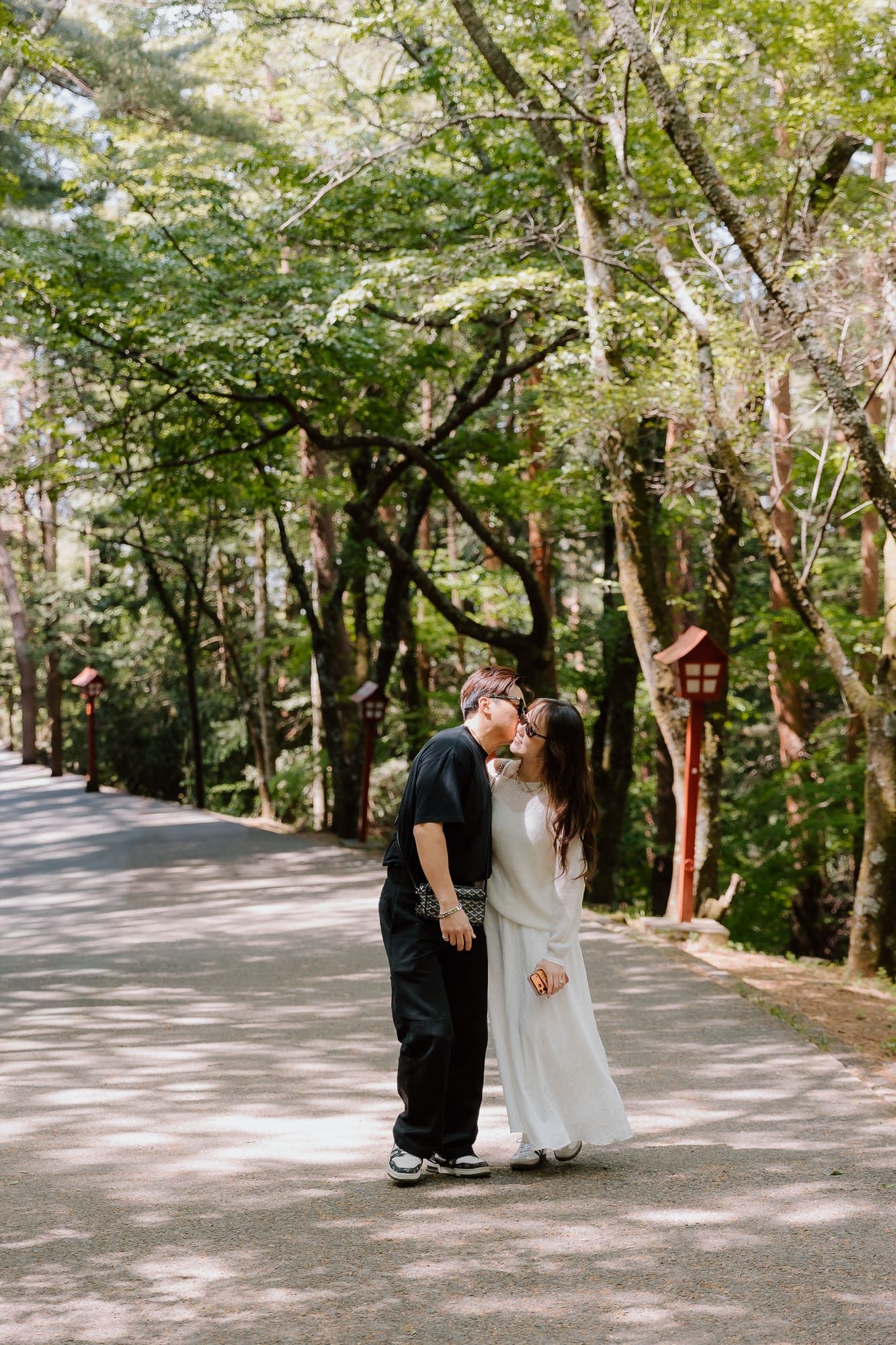 Couple sharing a romantic kiss on a tree-lined path in a lush Japanese forest setting.