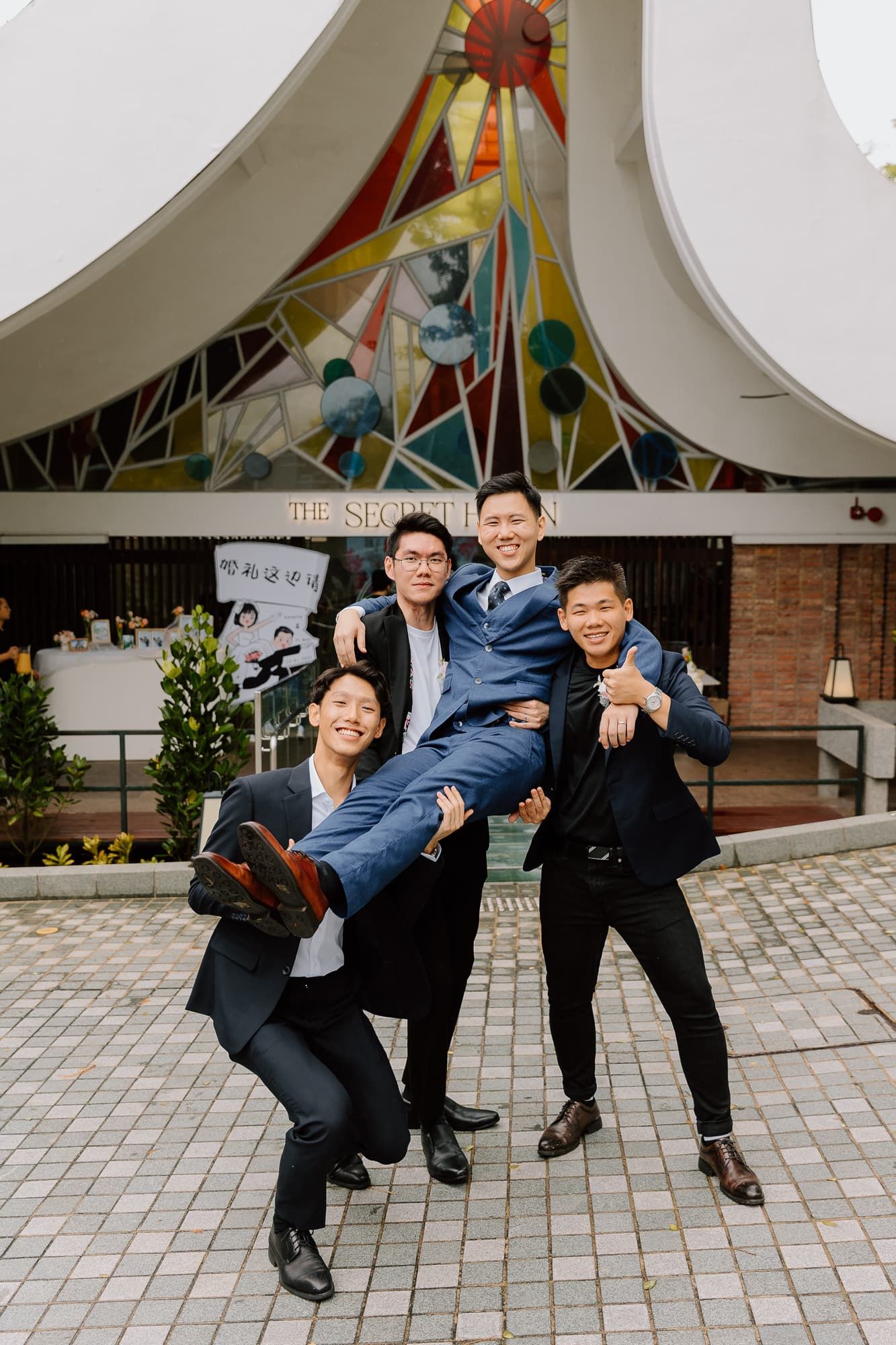 Playful groomsmen lifting and posing with groom under decorative ceiling.