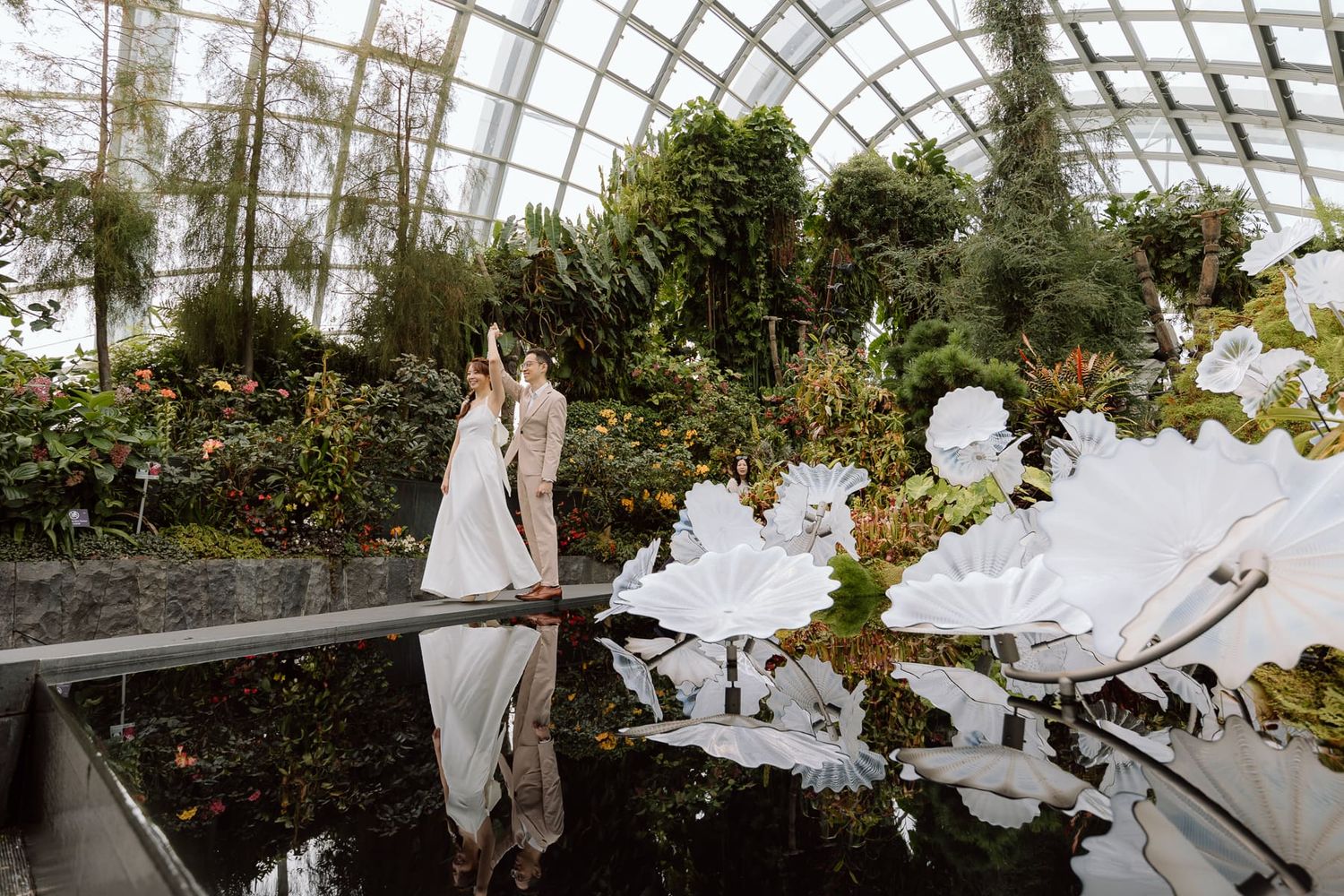 A couple reflected in still water at a greenhouse conservatory surrounded by lush greenery.