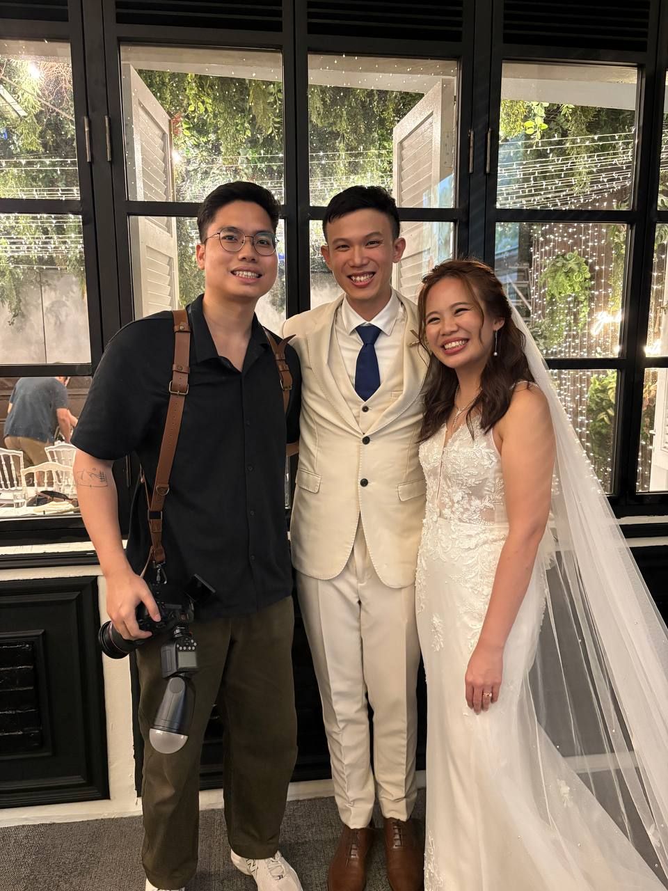Three people pose together at a wedding, with the newlyweds wearing white attire and a photographer in casual clothing.