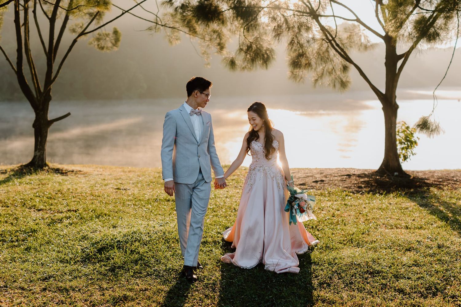 Couple walking together on grassy lakeside during golden hour photoshoot.