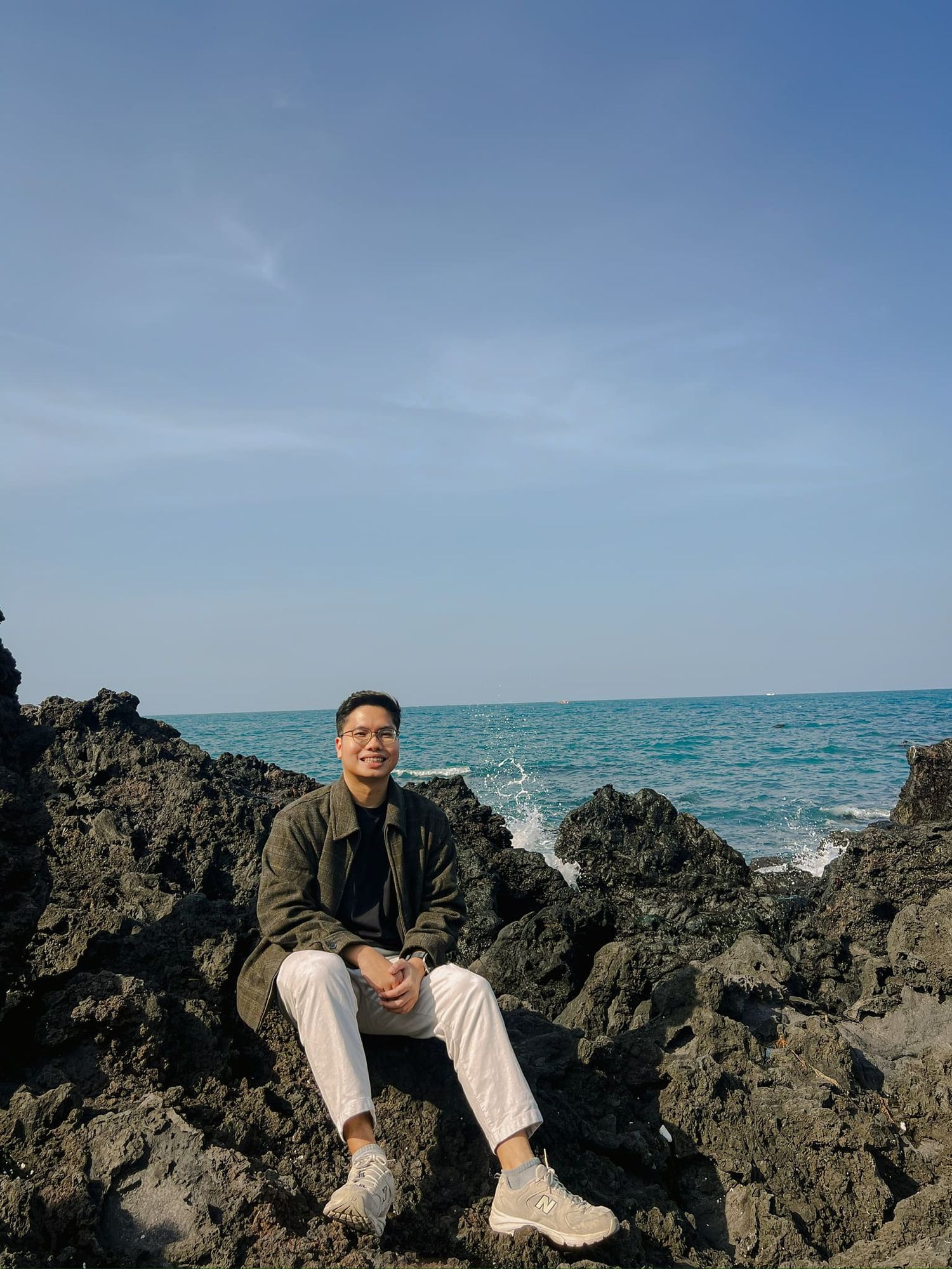 A young man smiling while sitting on rocky coastal cliffs with turquoise ocean waves and blue sky behind him.