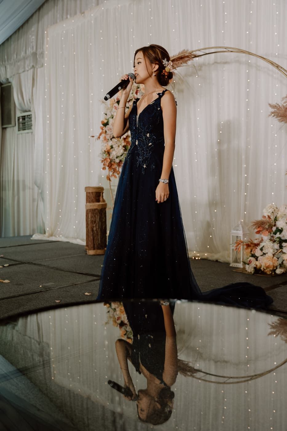 Person in a sparkling black gown performs with a microphone against a fairy light backdrop.