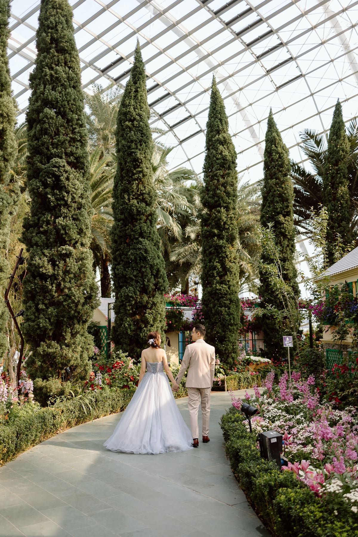A couple walks hand in hand through a conservatory garden path lined with tall cypress trees.