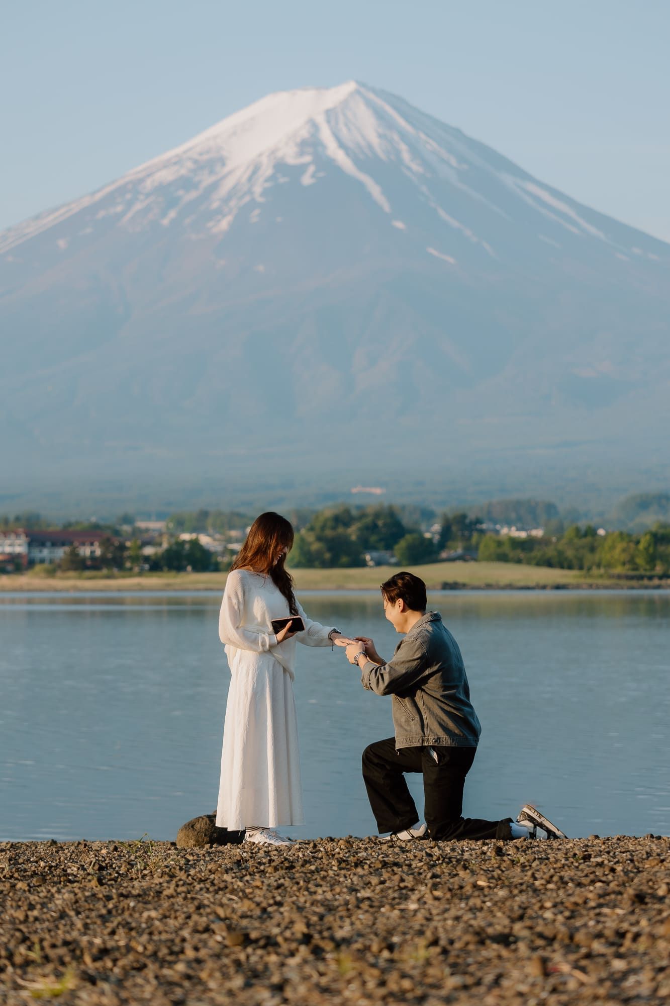 Man proposing to woman in white dress by lake with Mount Fuji backdrop.