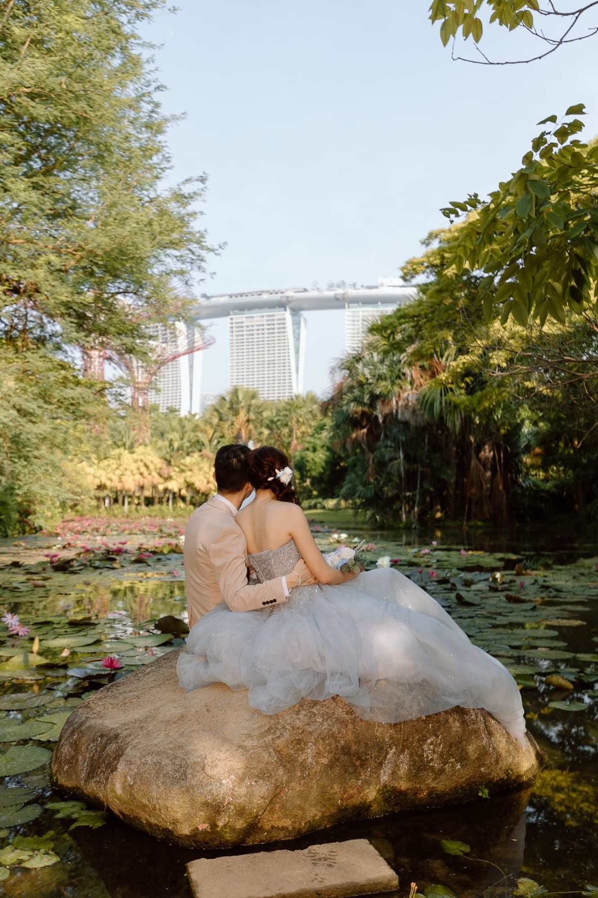 A wedding couple sitting on a rock by a lily pond with Marina Bay Sands in the distance.