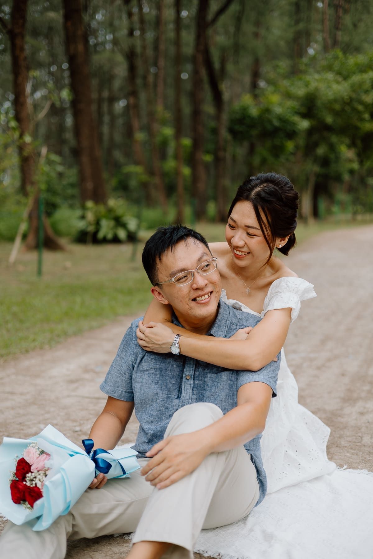 Playful couple sitting on sandy ground with flowers in a pine forest setting.