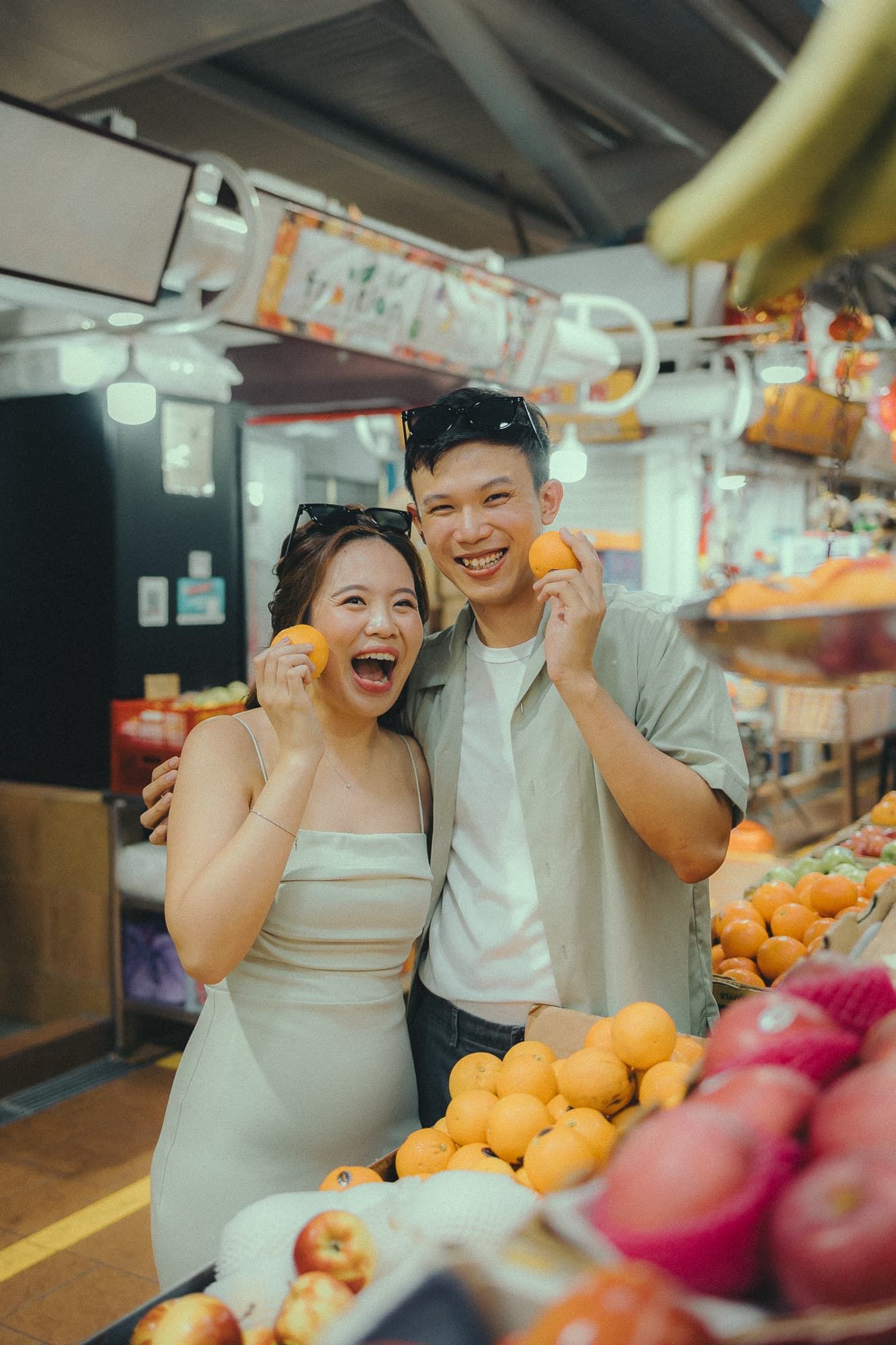 A joyful couple poses with oranges at a local market stall filled with fresh fruits.
