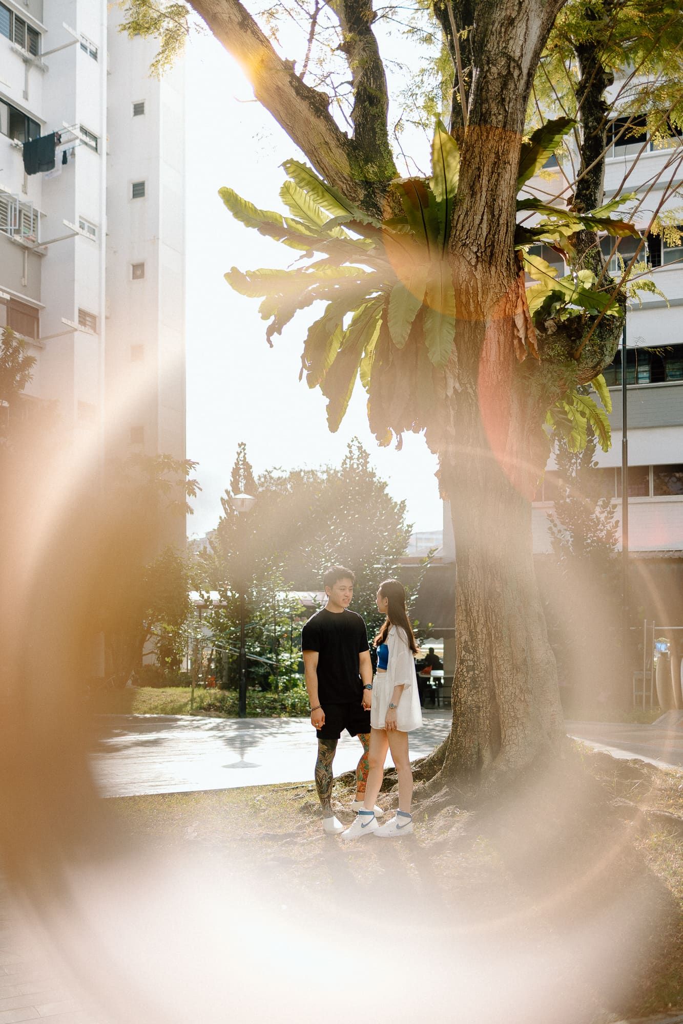 A couple walking together on a sunlit path with trees and apartment buildings in the background.