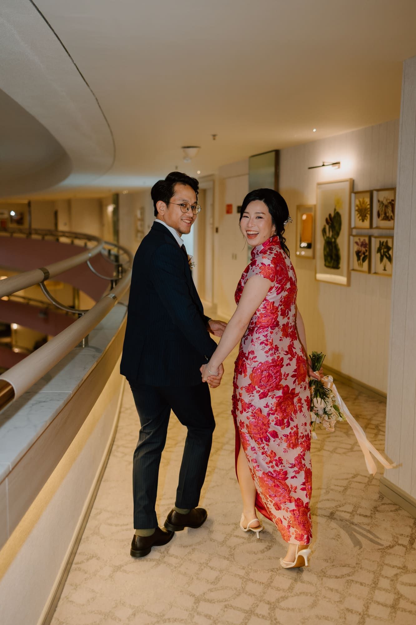 A couple poses in formal attire in a hotel hallway, with the woman wearing a traditional red floral qipao dress.