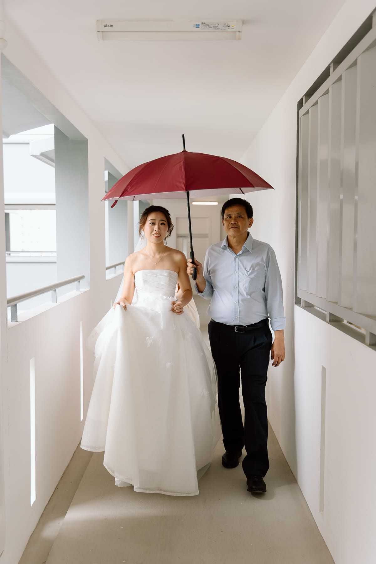 Wedding couple walks down hallway under red umbrella in simple modern setting.