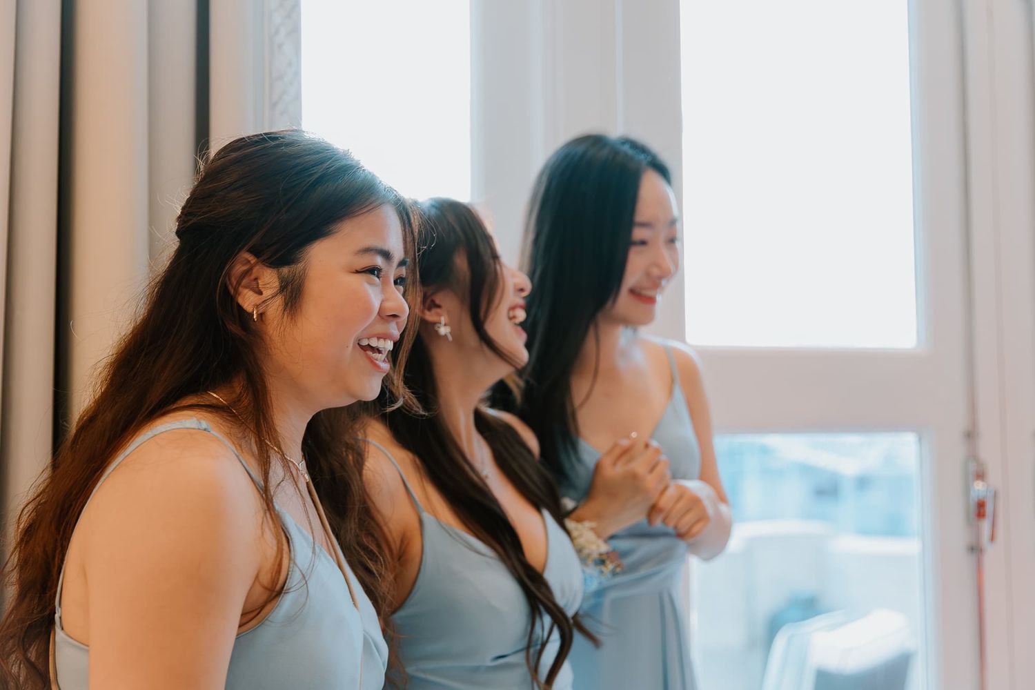 Bridesmaids in light blue dresses pose together by large windows in a modern room.