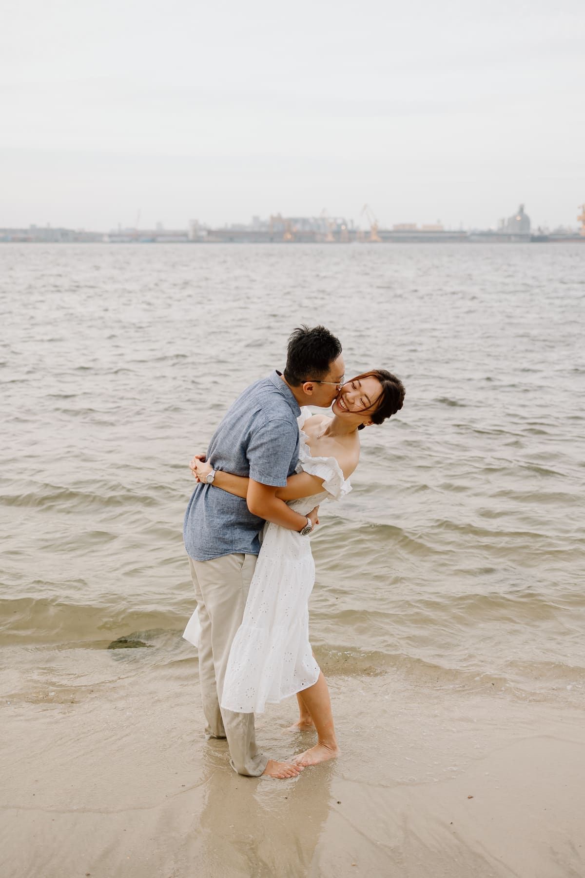A romantic couple embraces and kisses on a beach with a city skyline visible in the misty background.