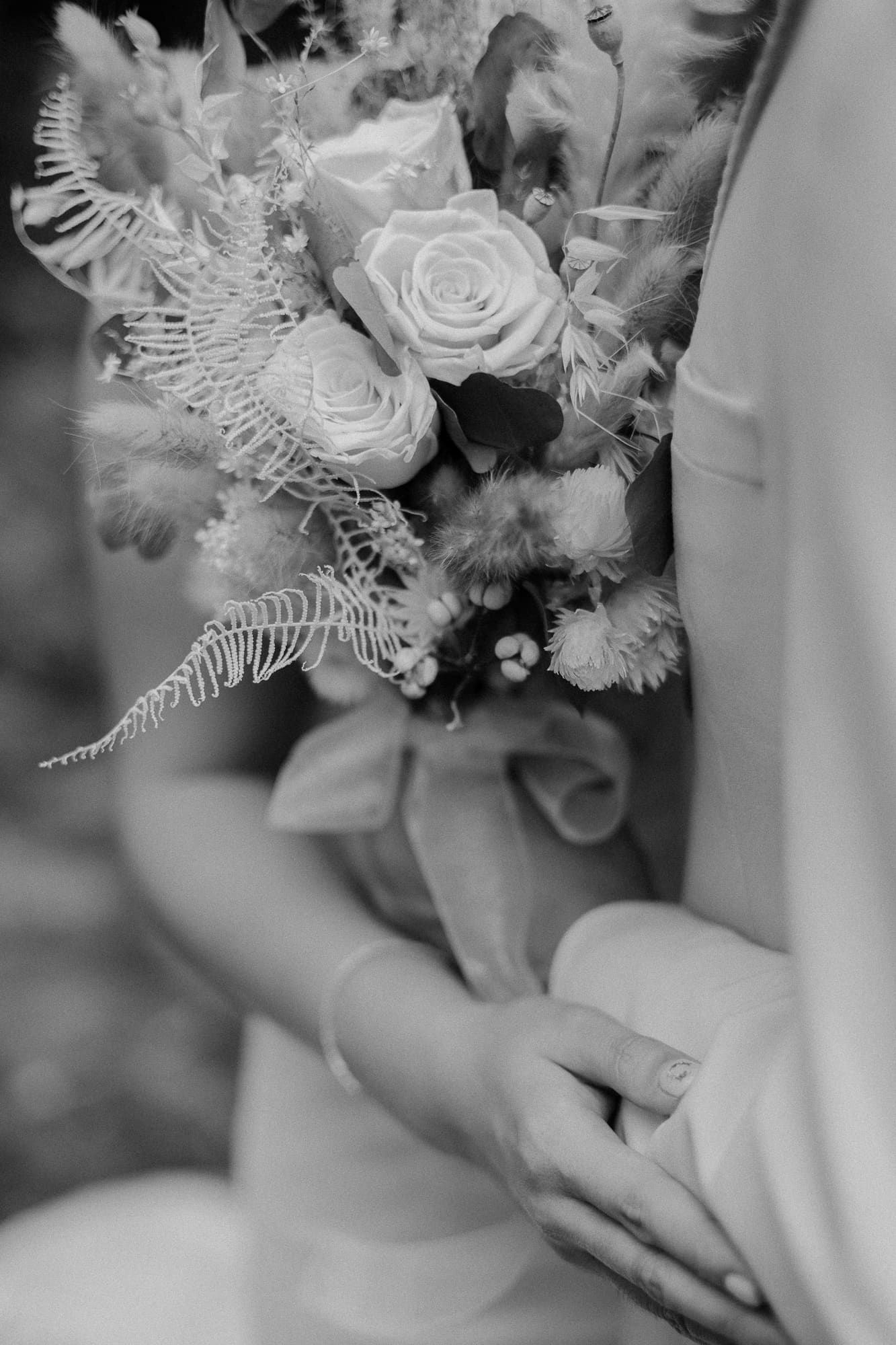 A close-up shot of elegant hands holding a delicate white rose and fern wedding bouquet.