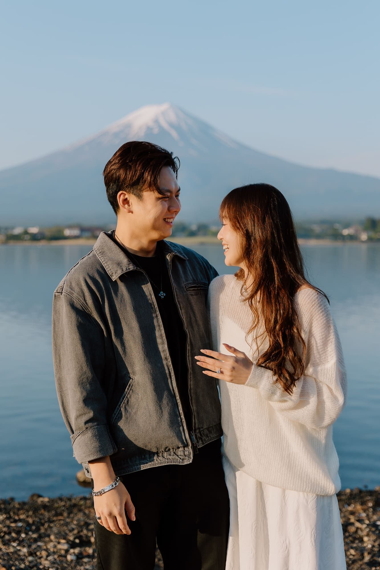 Couple smiling at each other near a lake with Mount Fuji in the background on a clear day.