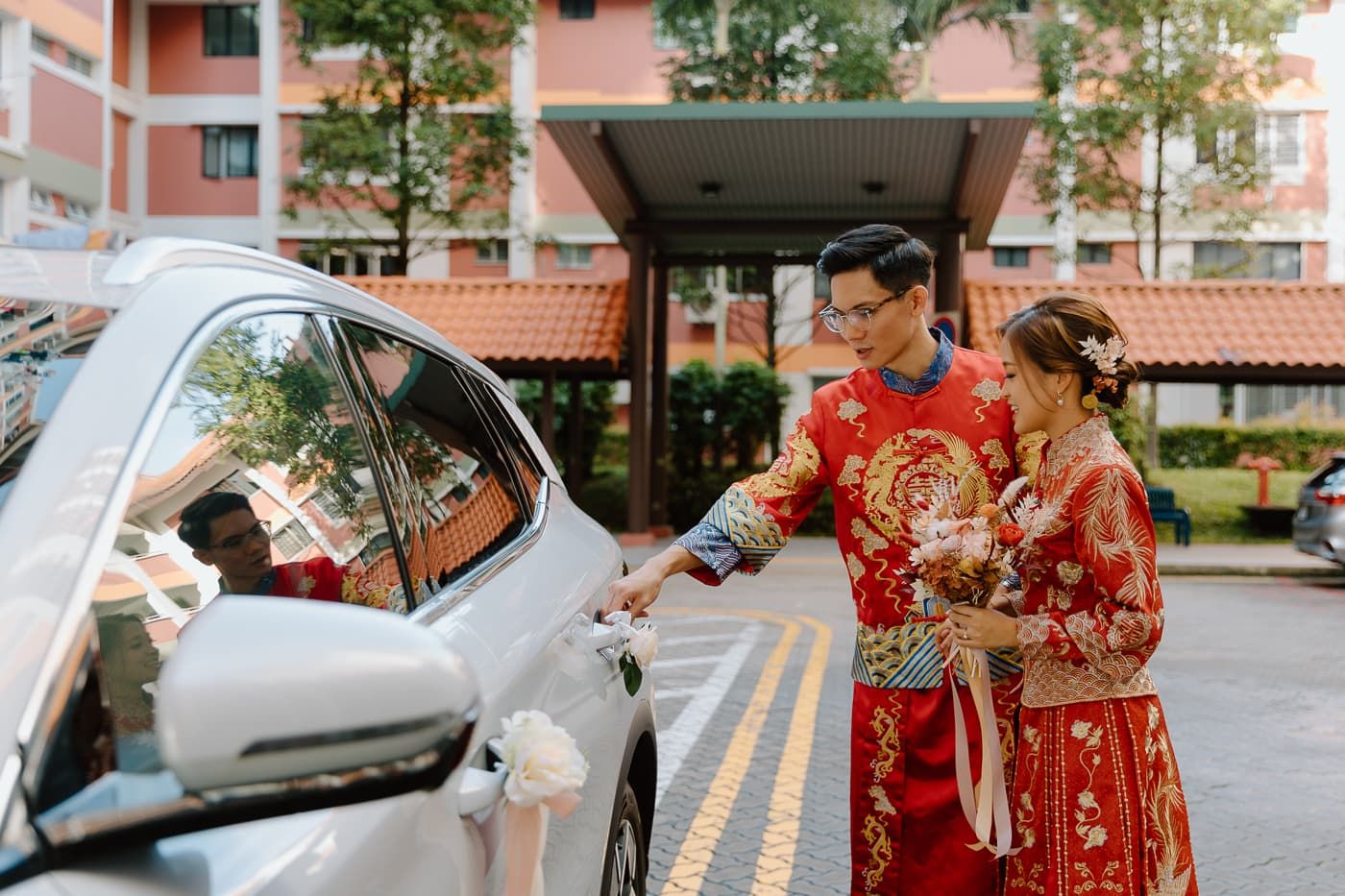 Couple in traditional red Chinese wedding attire greeting guests by their car outside a building.