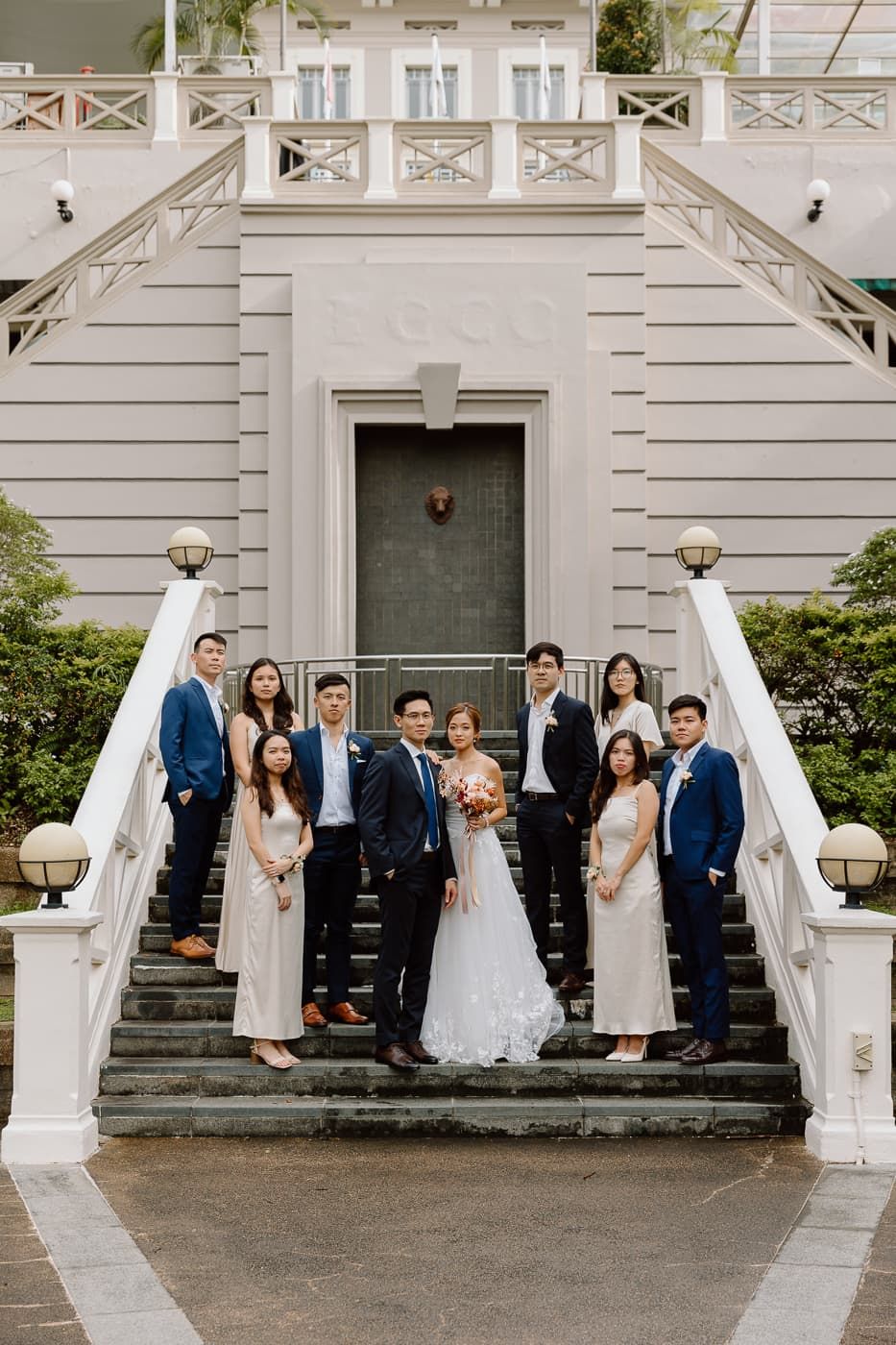 Wedding party poses on grand white staircase in front of historic building with green door and white railings.