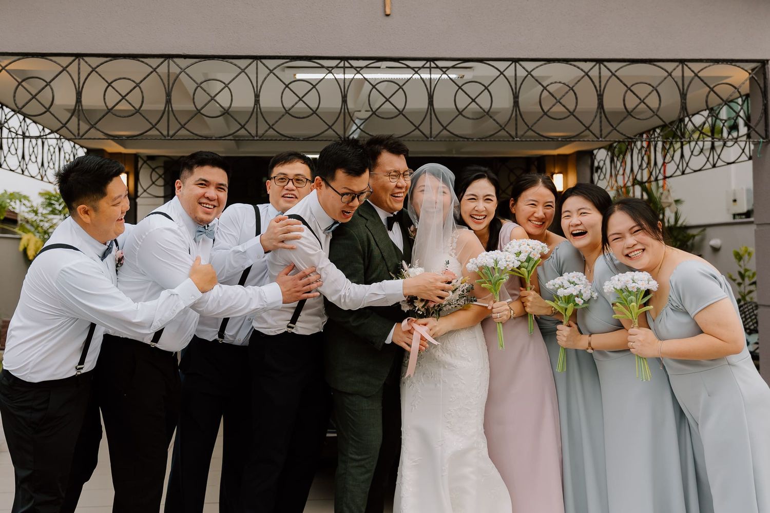 Wedding party members in gray suits and pastel dresses pose together in a fun group shot.