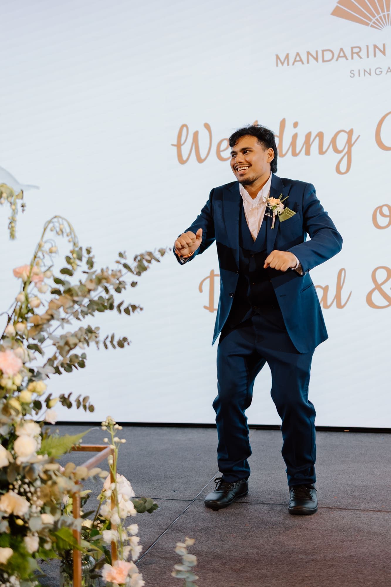 Person in navy blue suit crouches down during a wedding celebration with decorative backdrop visible.