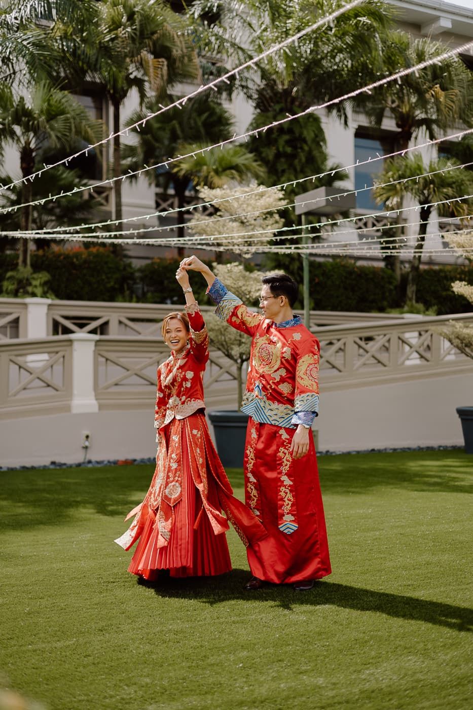 Couple in red traditional Chinese wedding attire dancing together on garden lawn.