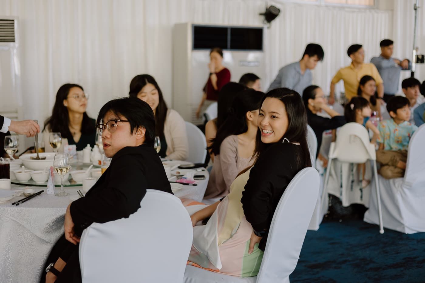 Wedding reception guests seated at decorated tables in a bright white venue.