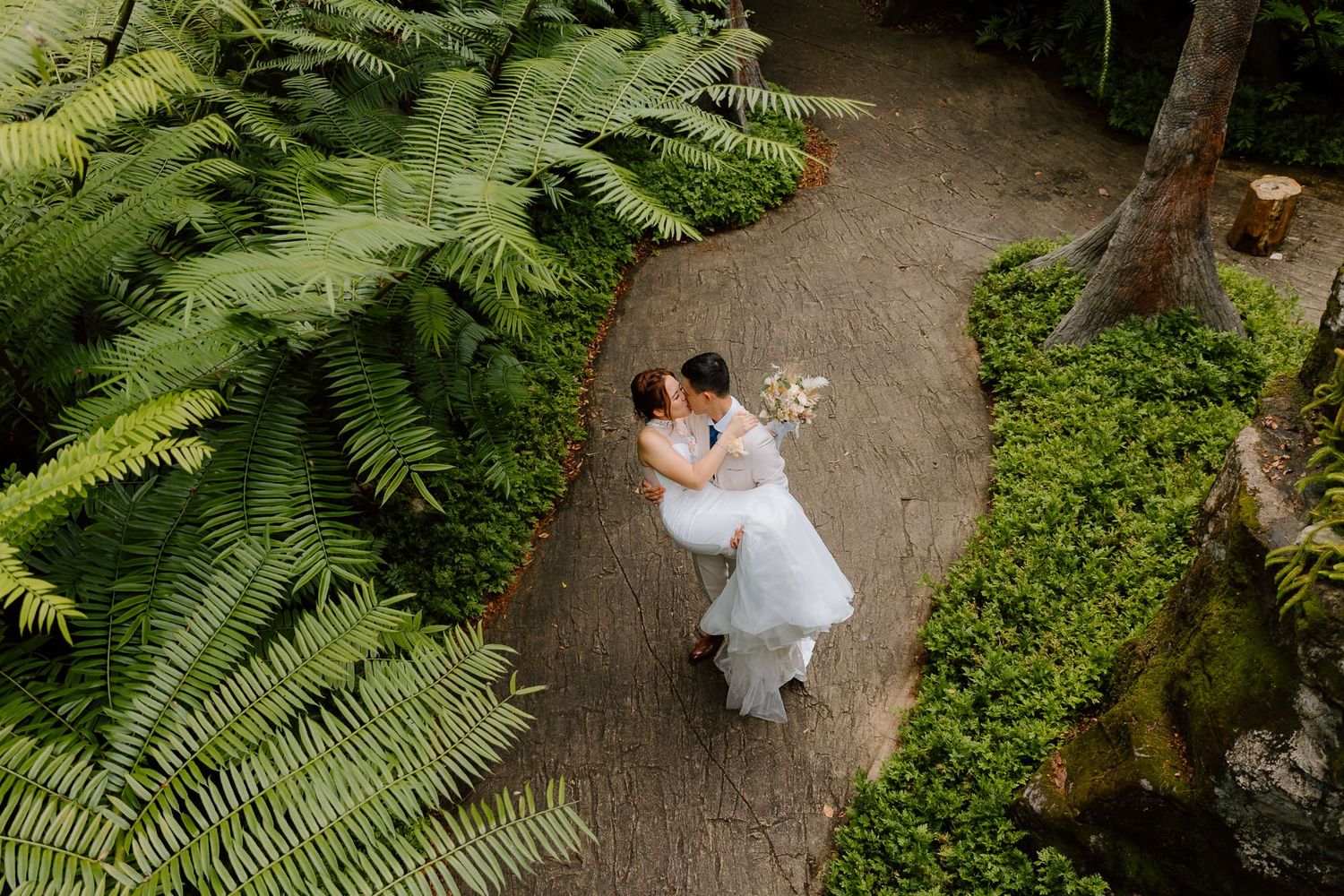 A couple walks hand in hand along a lush garden path surrounded by vibrant green ferns and foliage.