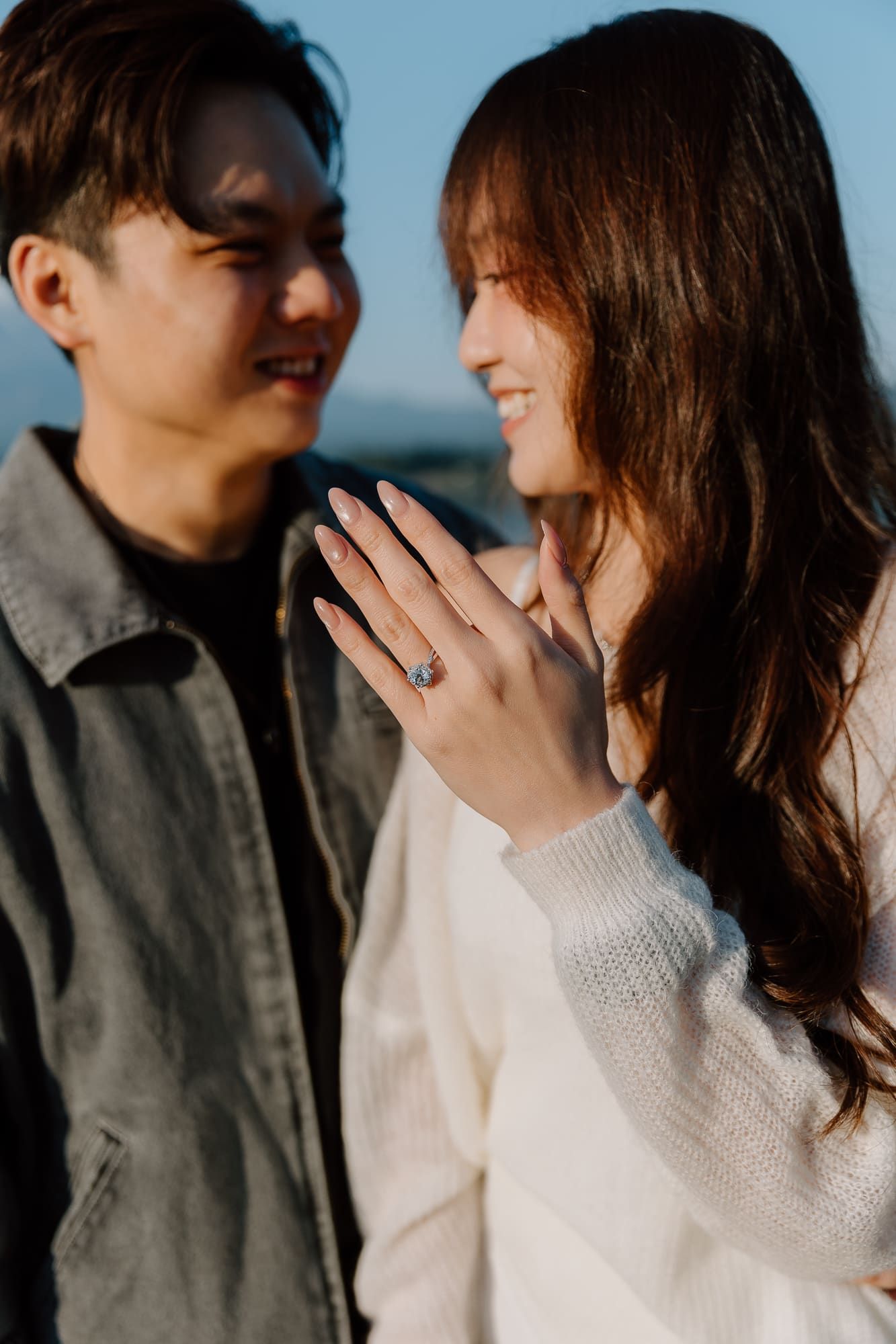 Smiling couple sharing a joyful moment as woman displays her diamond engagement ring in warm light.