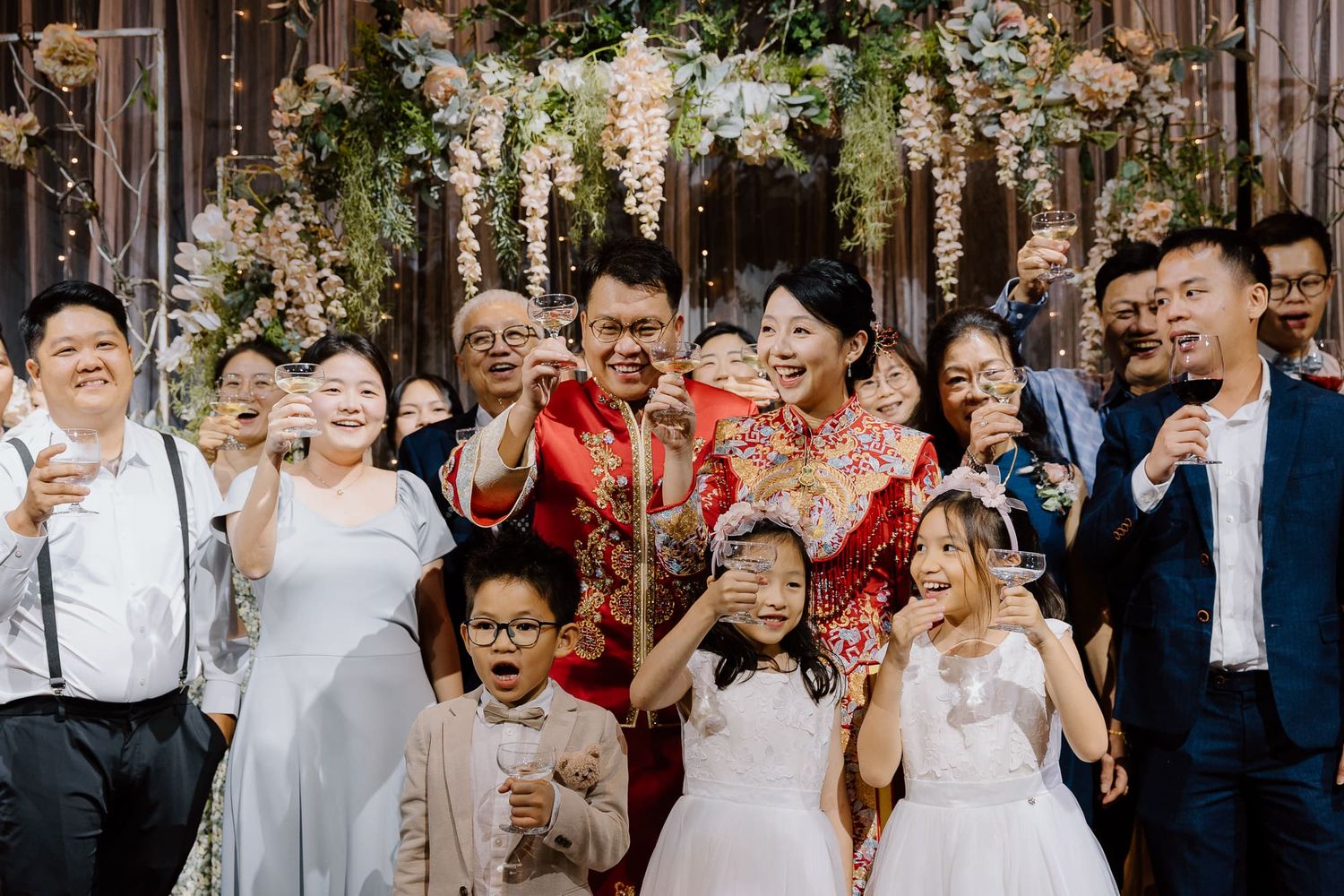 Wedding party celebrates joyfully under hanging white floral decorations at indoor ceremony venue.