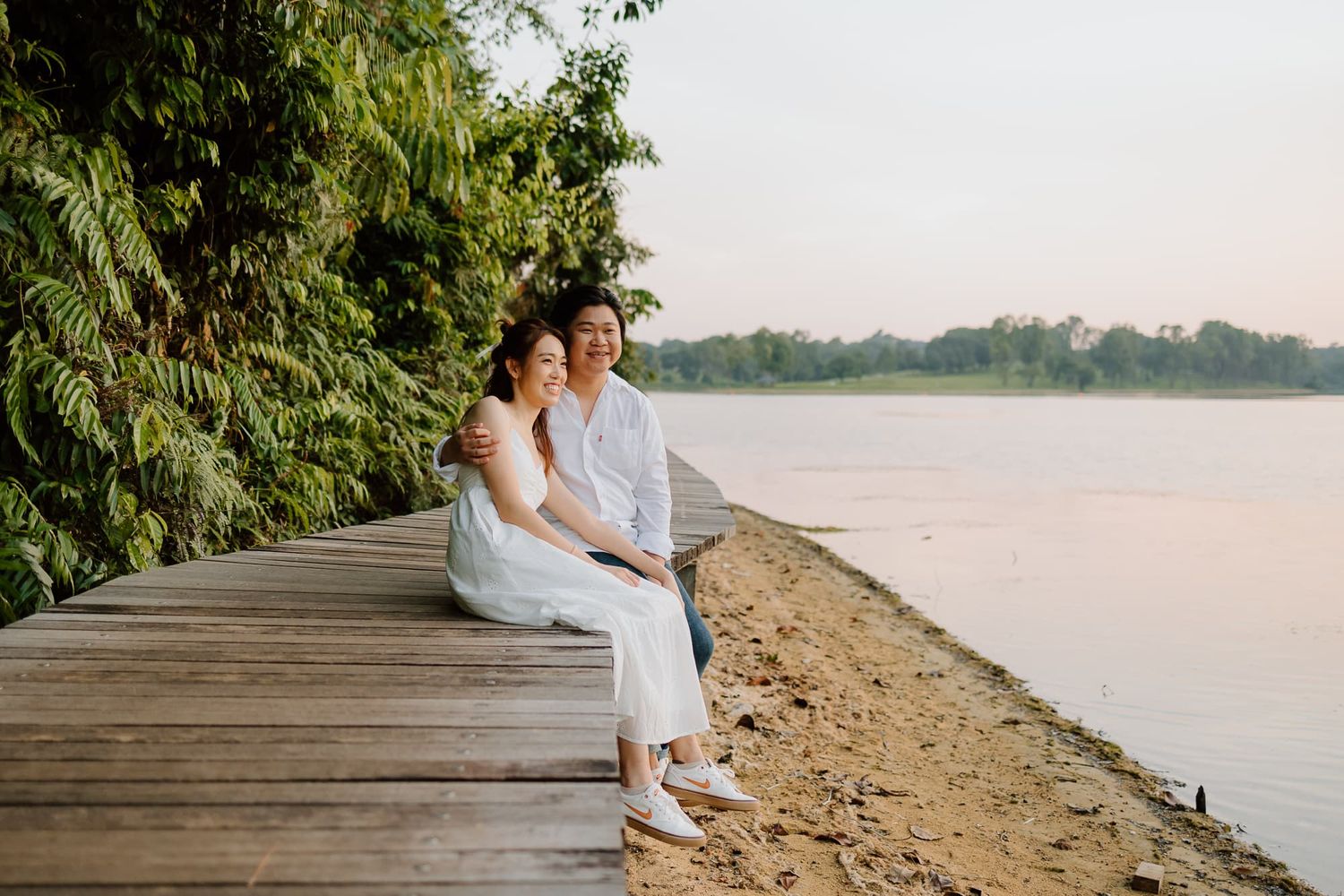Romantic couple sitting on wooden boardwalk by lake at sunset in white outfits.