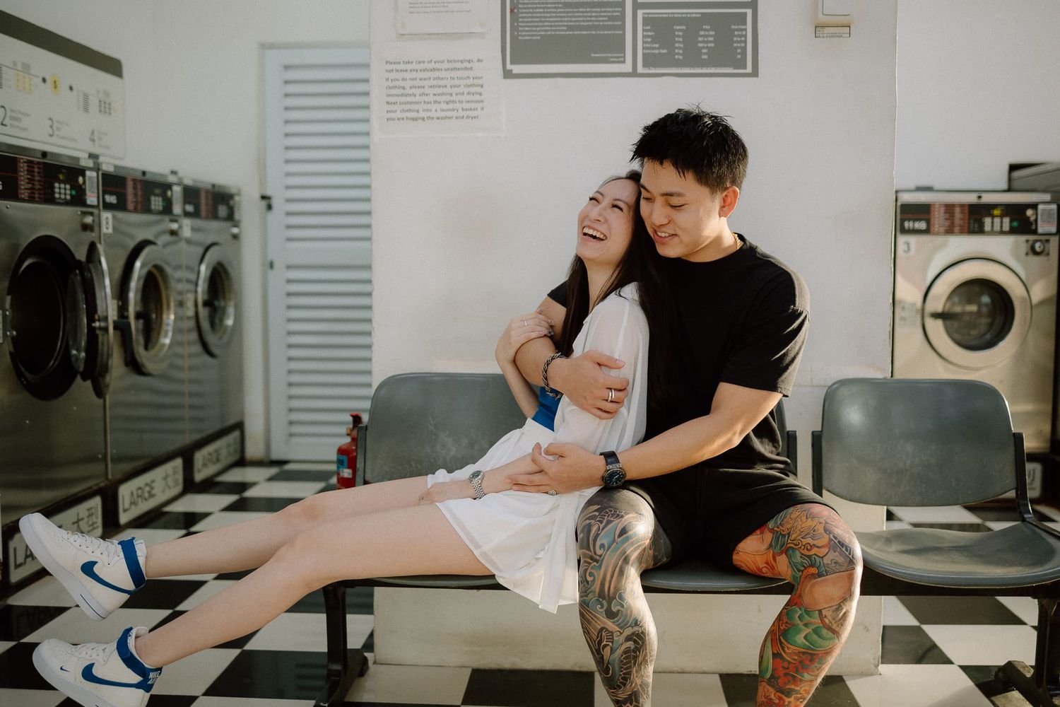 A romantic couple embraces and poses playfully in a laundromat setting with black and white checkered floors.