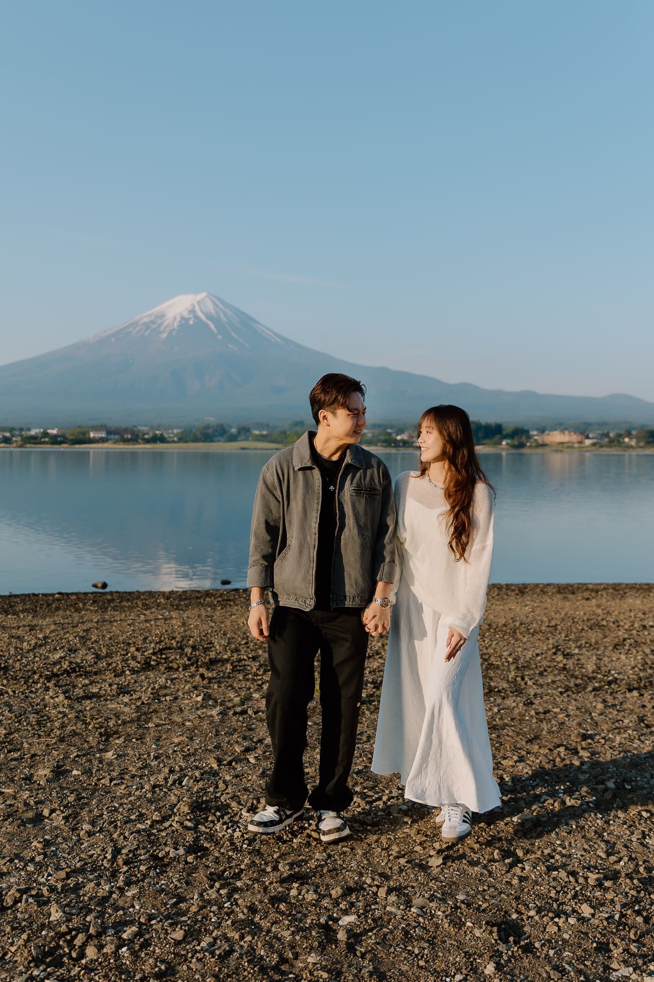 Couple holding hands by Lake Kawaguchi with iconic snow-capped Mount Fuji reflected in calm waters behind them.