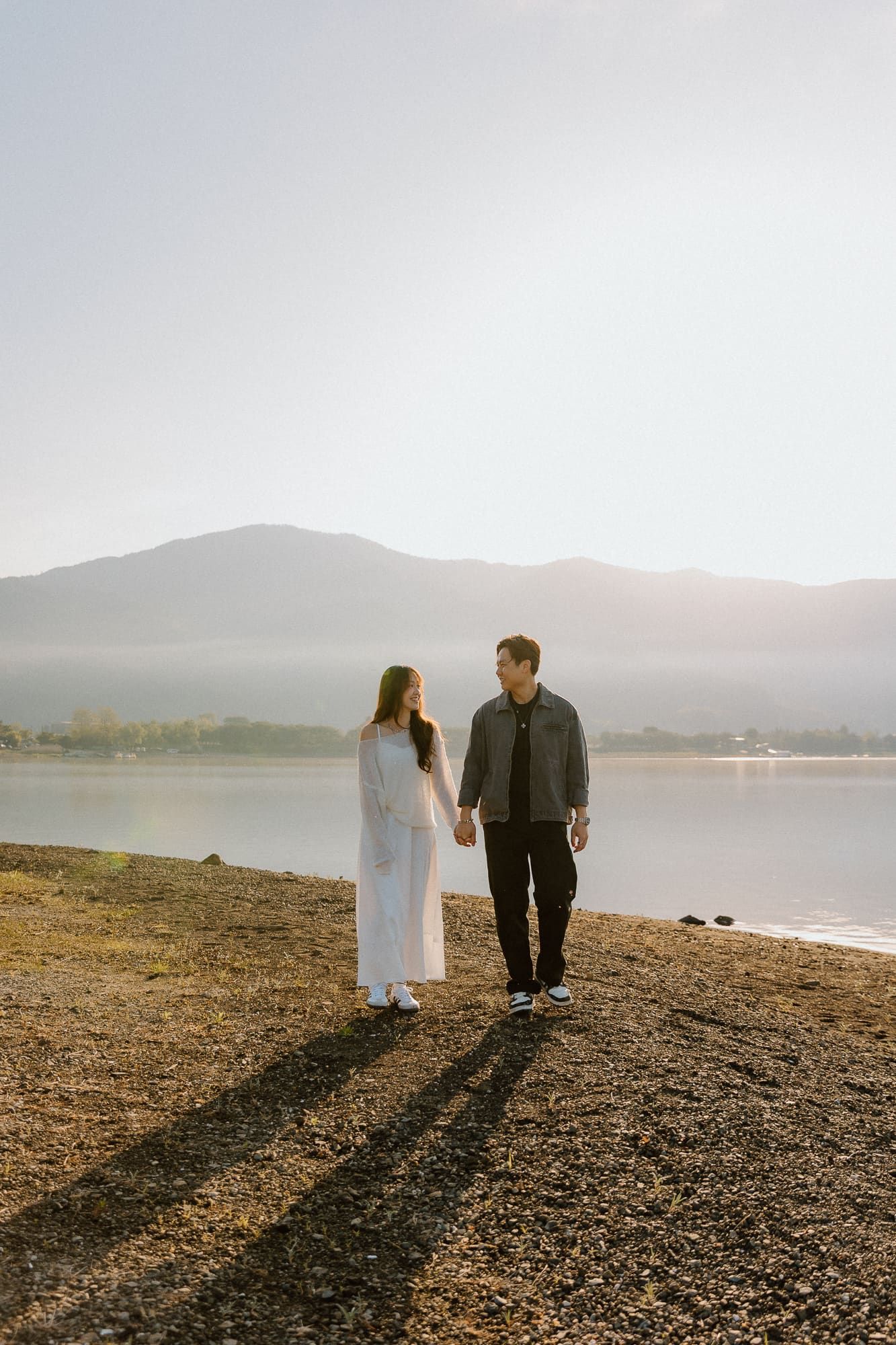 Happy couple walking hand in hand along a rocky lakeside with scenic mountain views at golden hour.