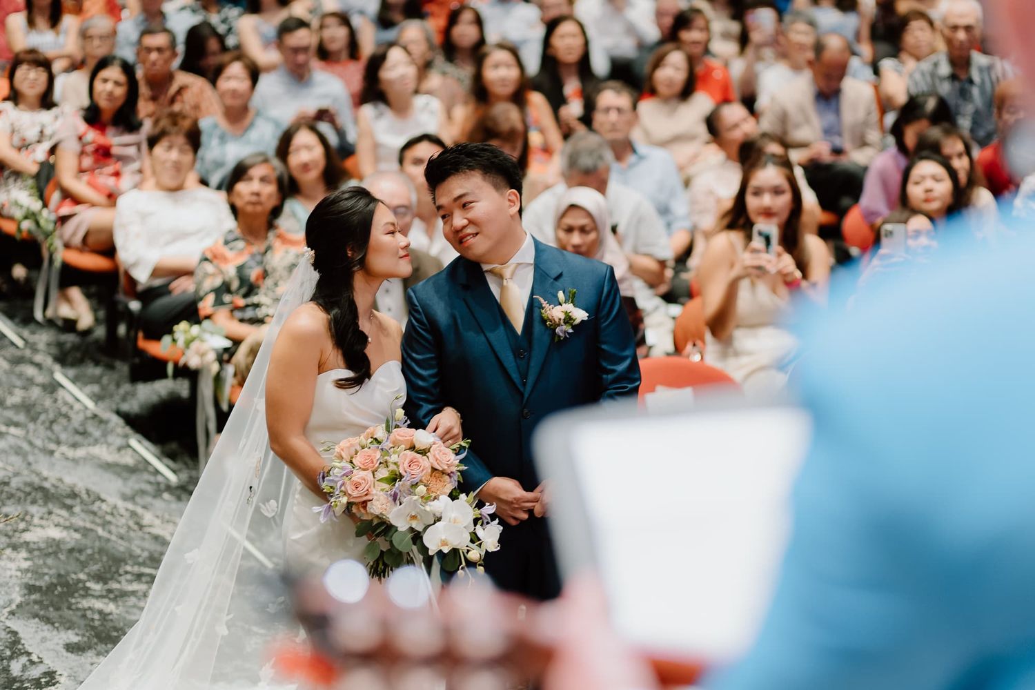 Wedding guests fill seats in a large indoor venue during a ceremony.