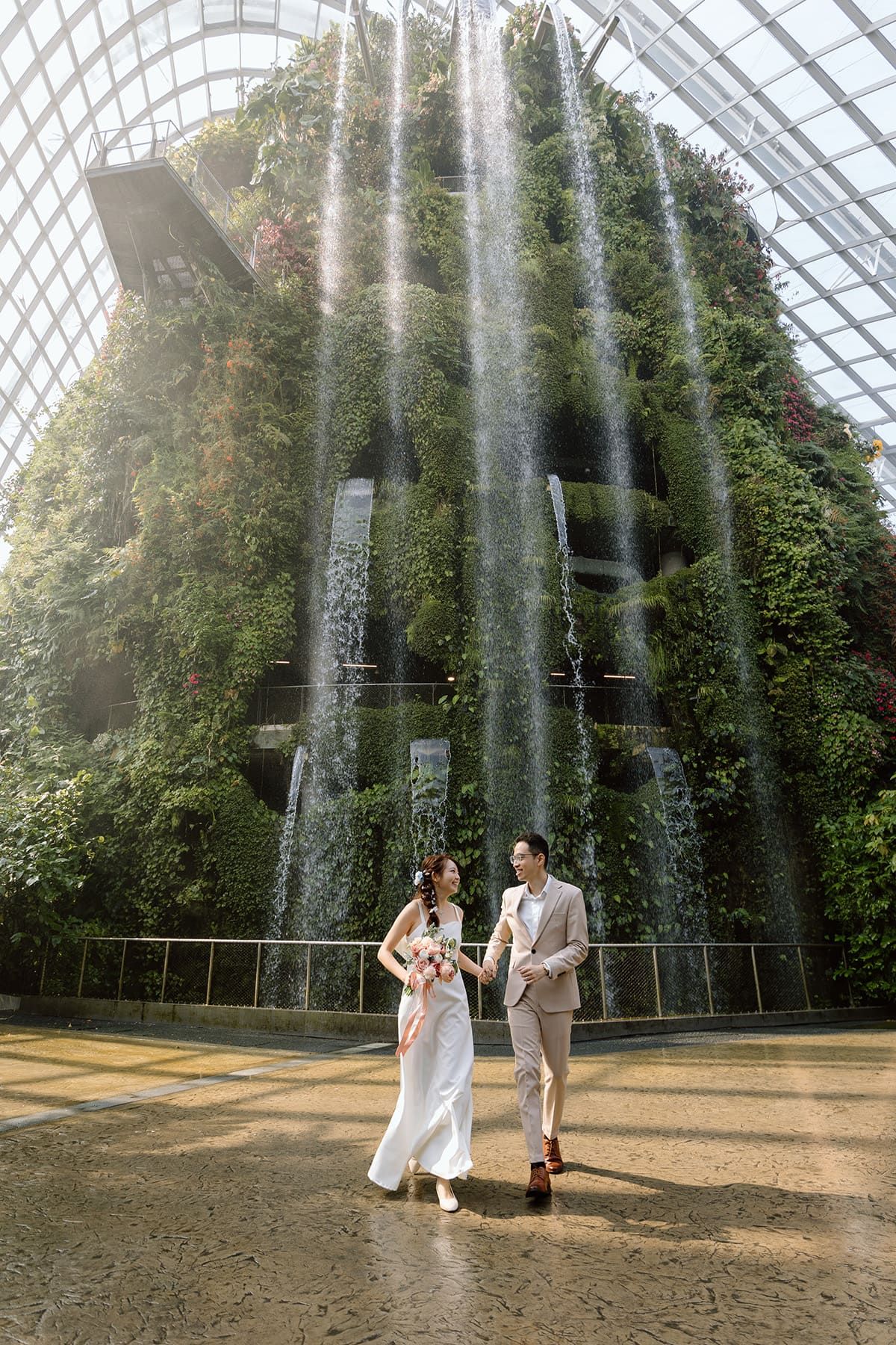 A dramatic wedding photo taken beneath a towering indoor waterfall in a glass-domed conservatory.