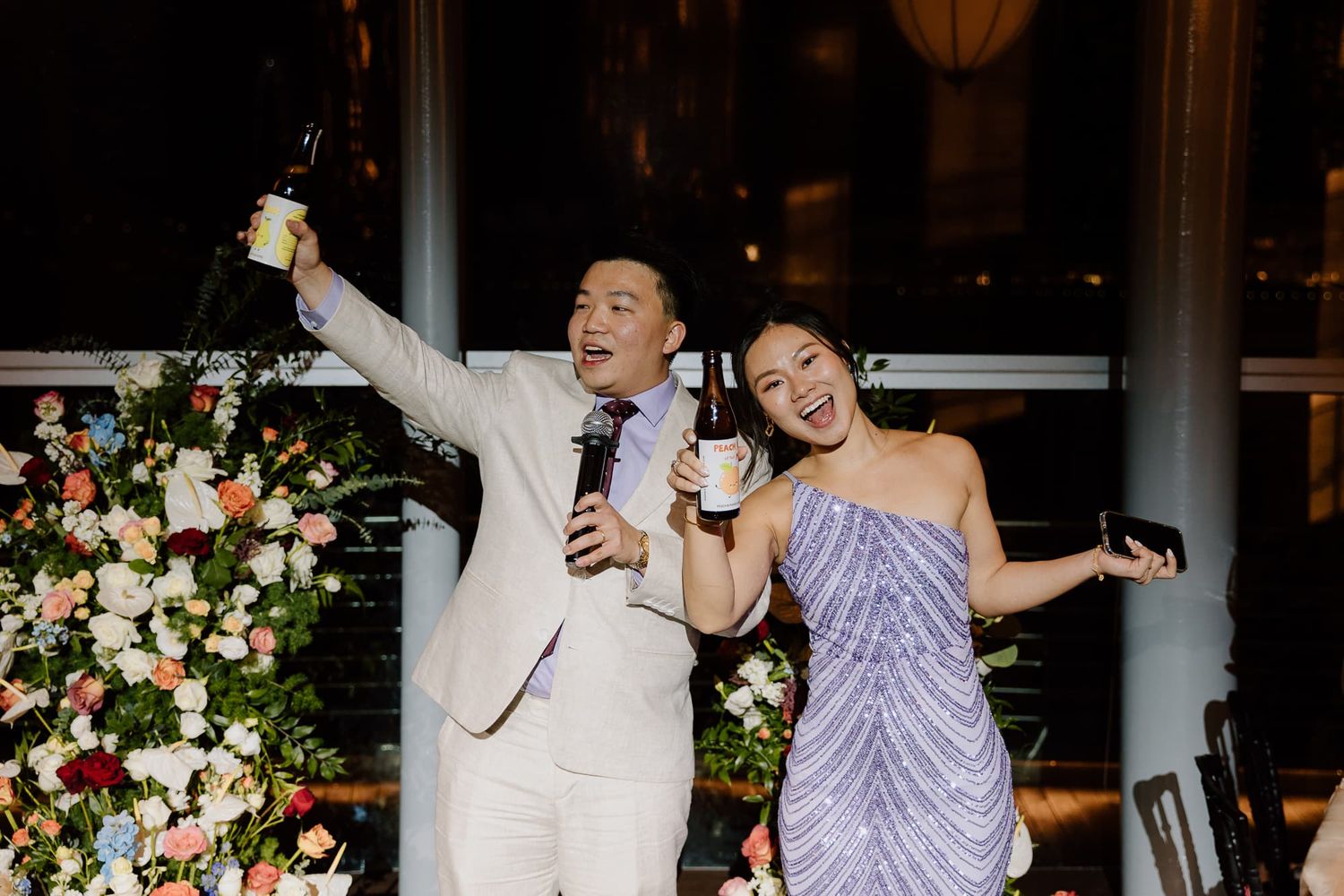 A couple shares a celebratory toast in front of a floral arrangement at an elegant evening reception.