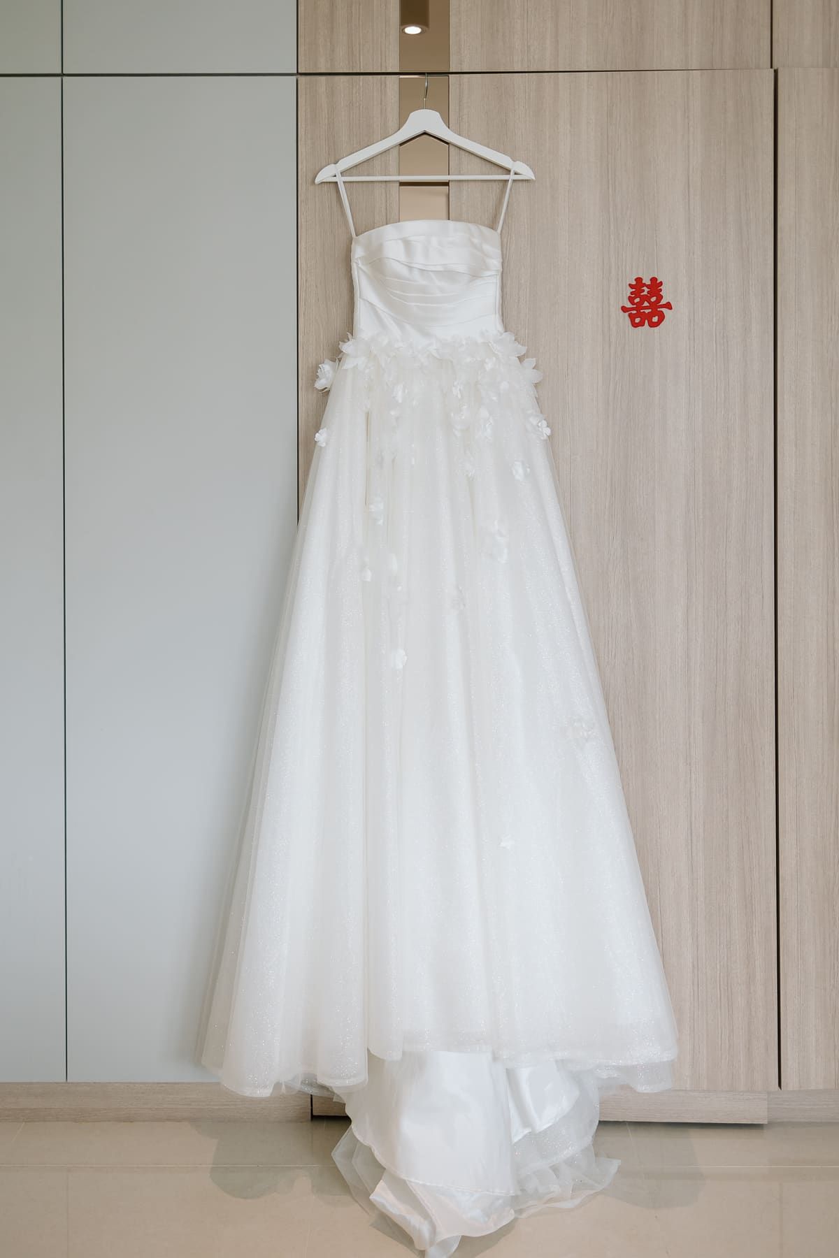An elegant white wedding dress with floral details hanging on display against a wooden wardrobe.