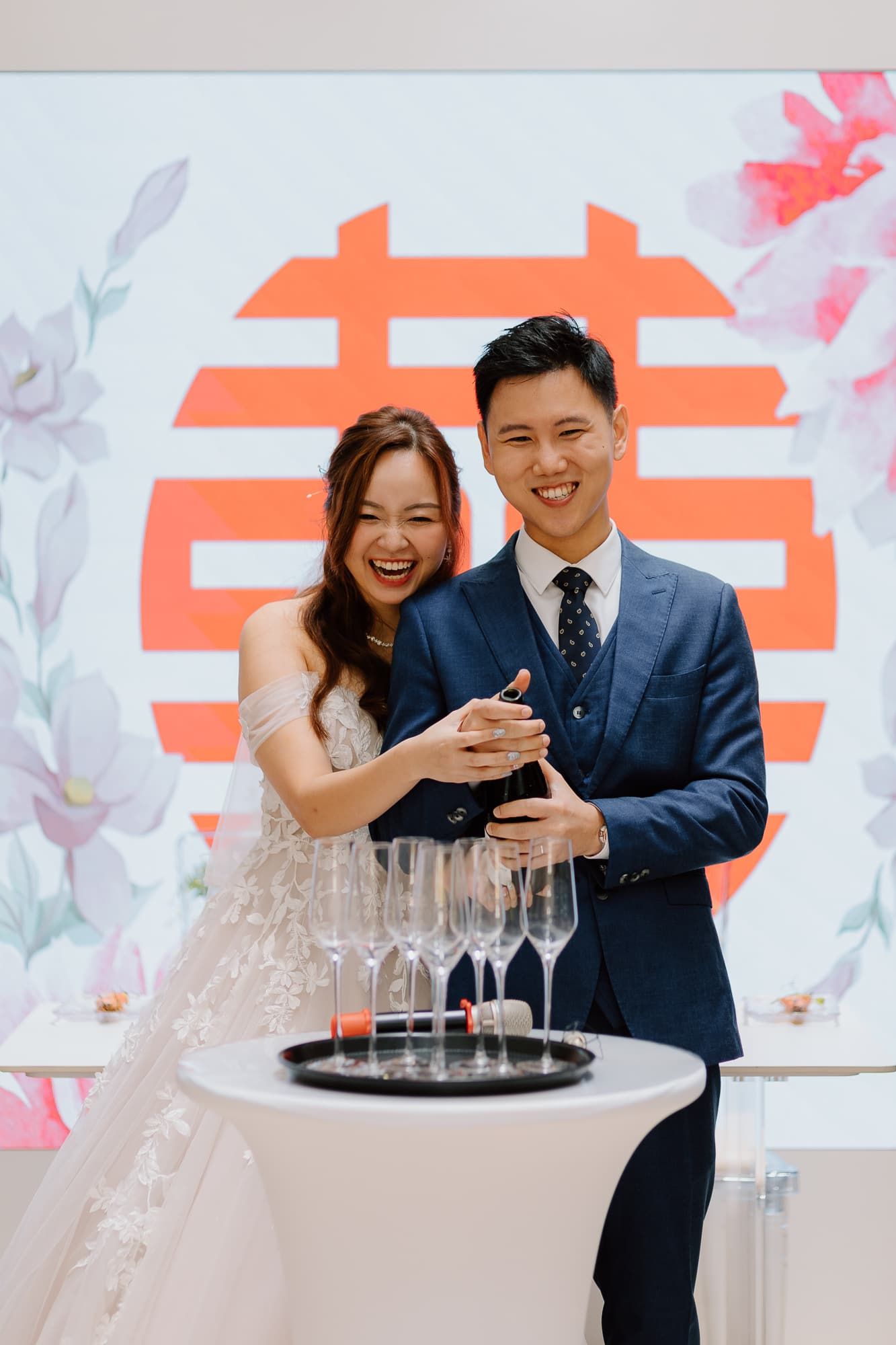 Couple sharing a champagne toast in front of traditional Chinese wedding backdrop with floral designs.