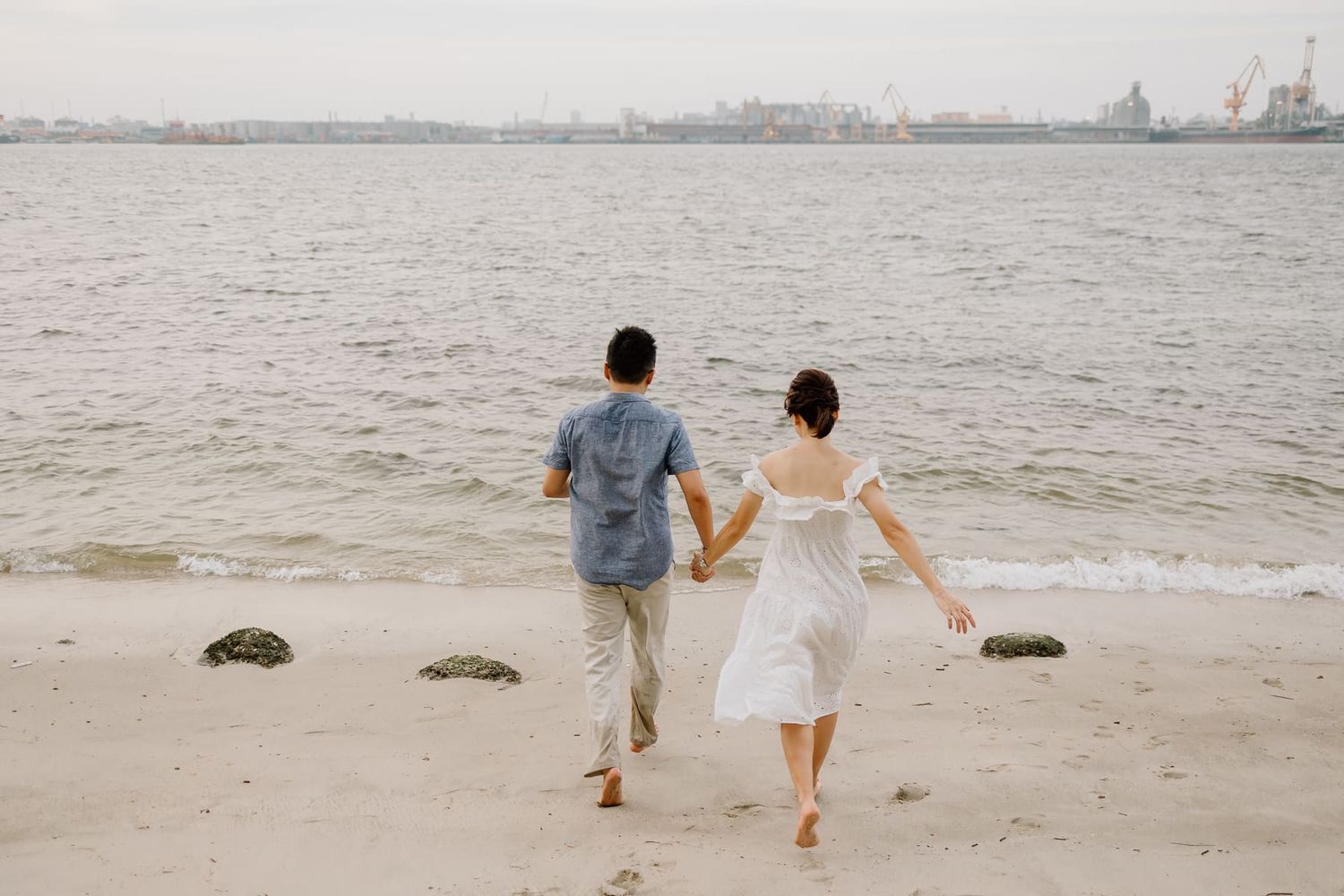 A couple walking hand in hand on a beach with city skyline in the distance.