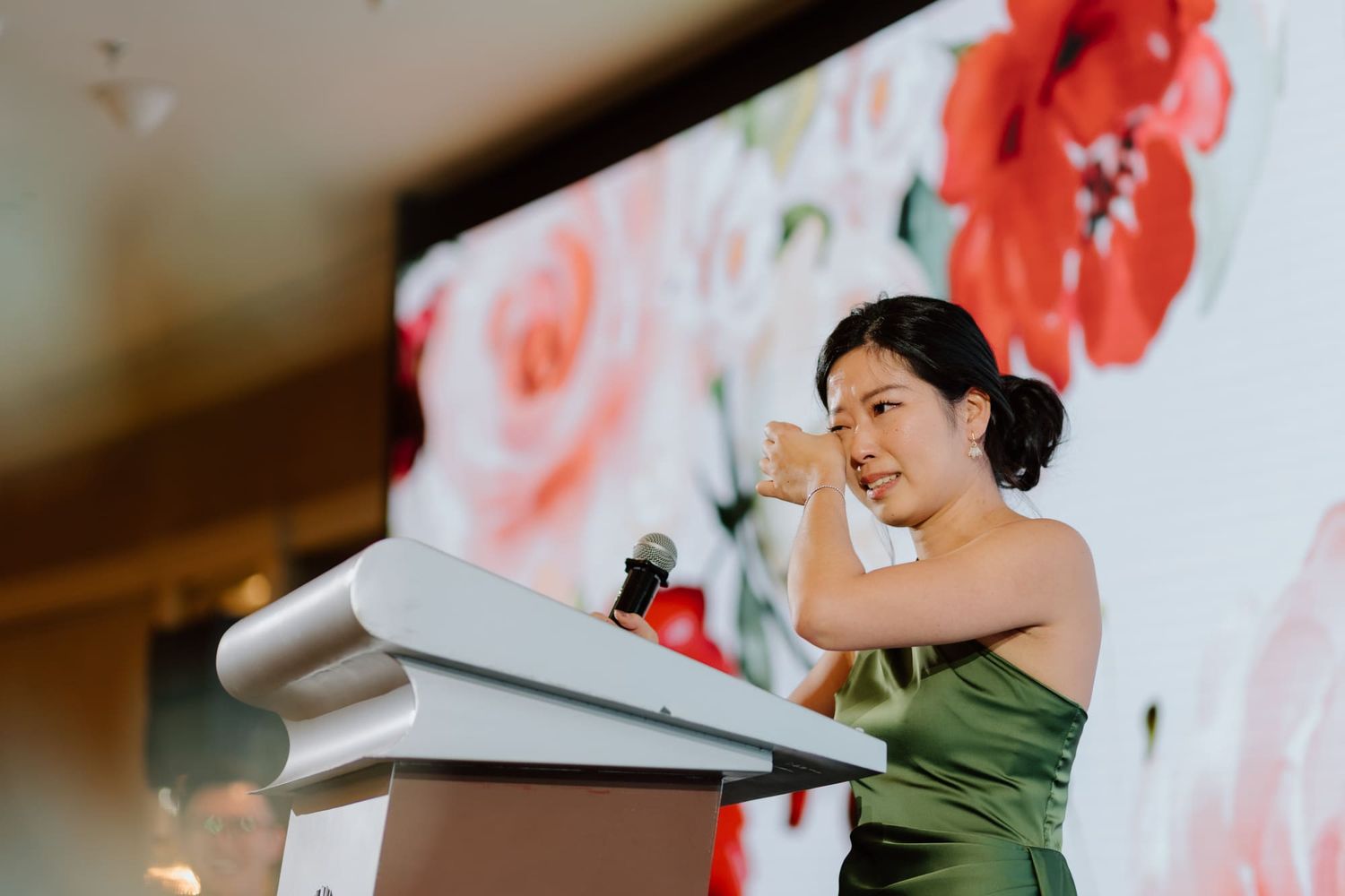 Speaker in emerald green dress delivers speech at podium with floral backdrop at formal event.