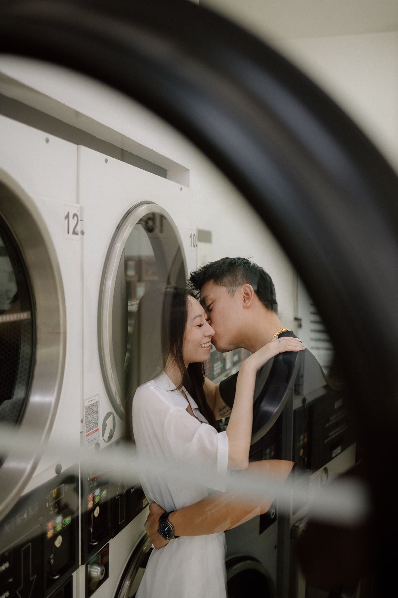 A romantic couple embraces in a laundromat between washing machines, creating an intimate moment in an everyday setting.