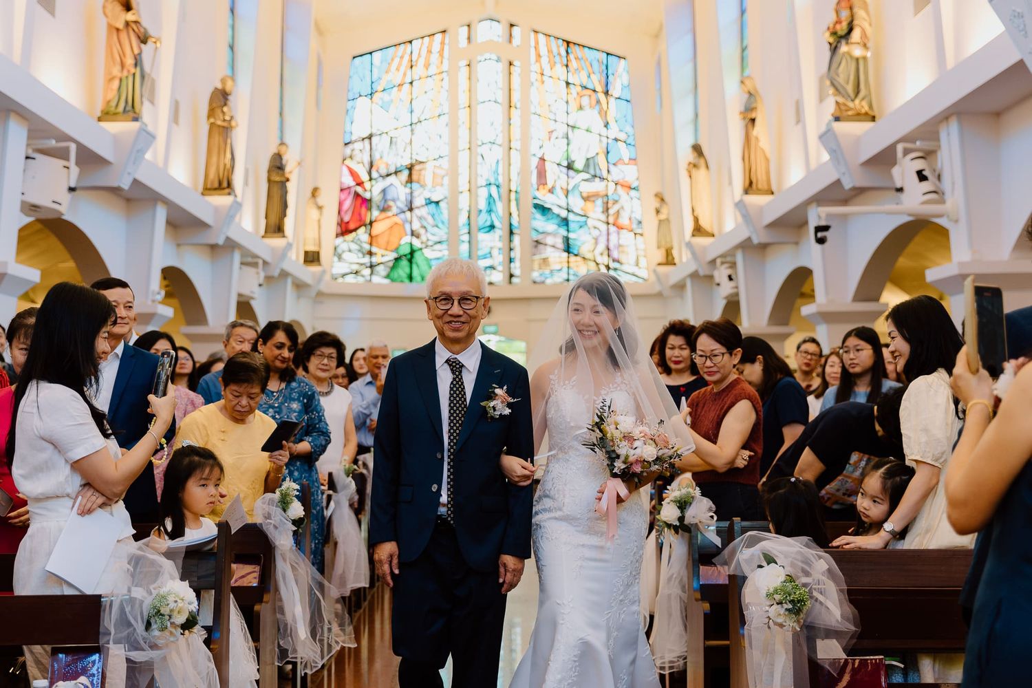 Wedding guests gather in an ornate white church with stained glass windows during a joyful ceremony celebration.