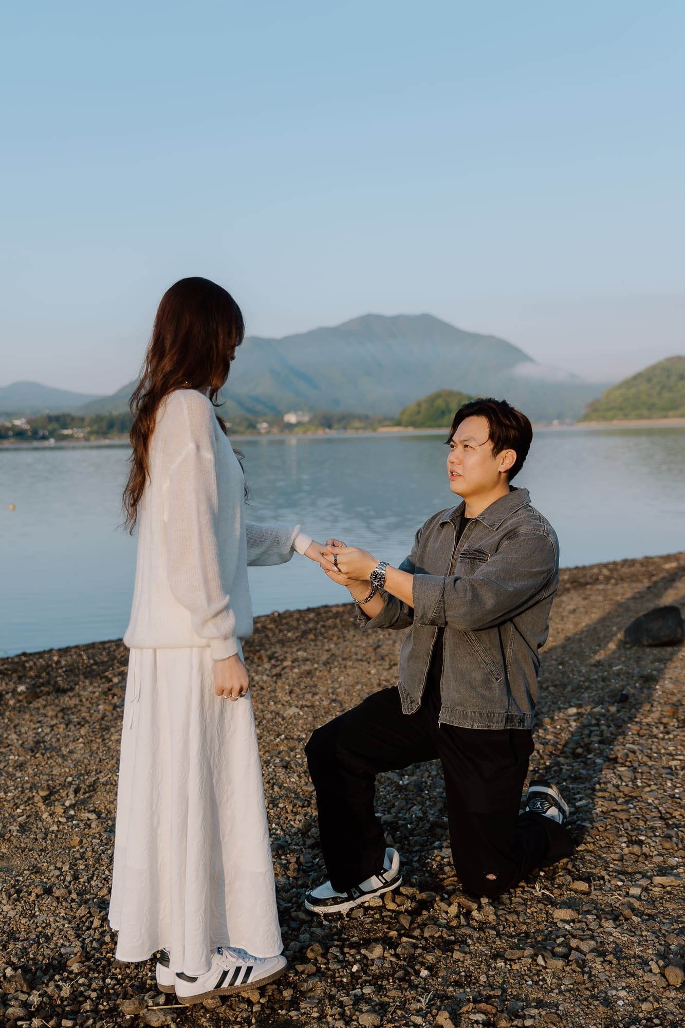 Man proposing to woman on one knee by a scenic lakeside with mountains in the background at dusk.