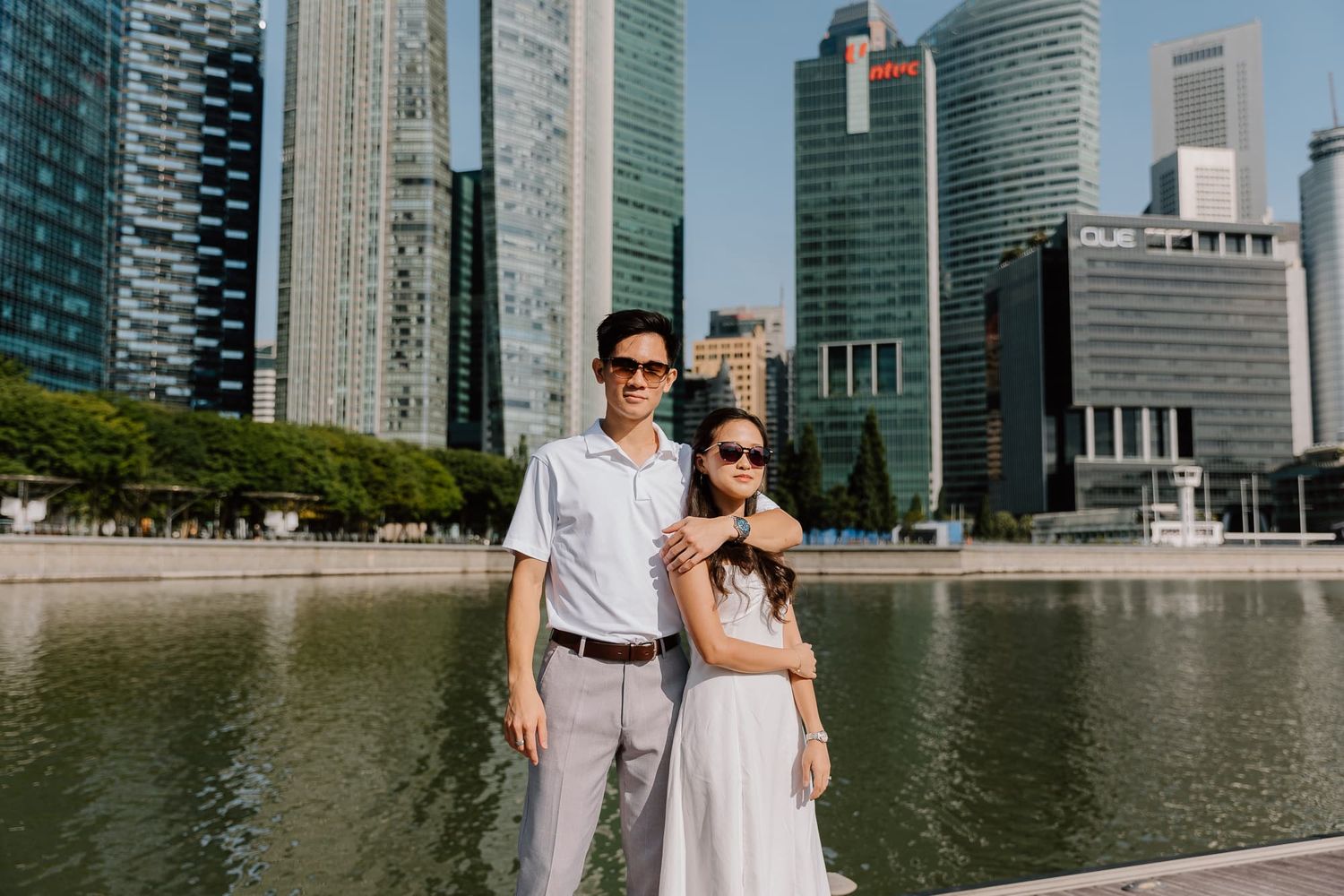 Couple in casual attire posing by waterfront with modern city skyline in background.