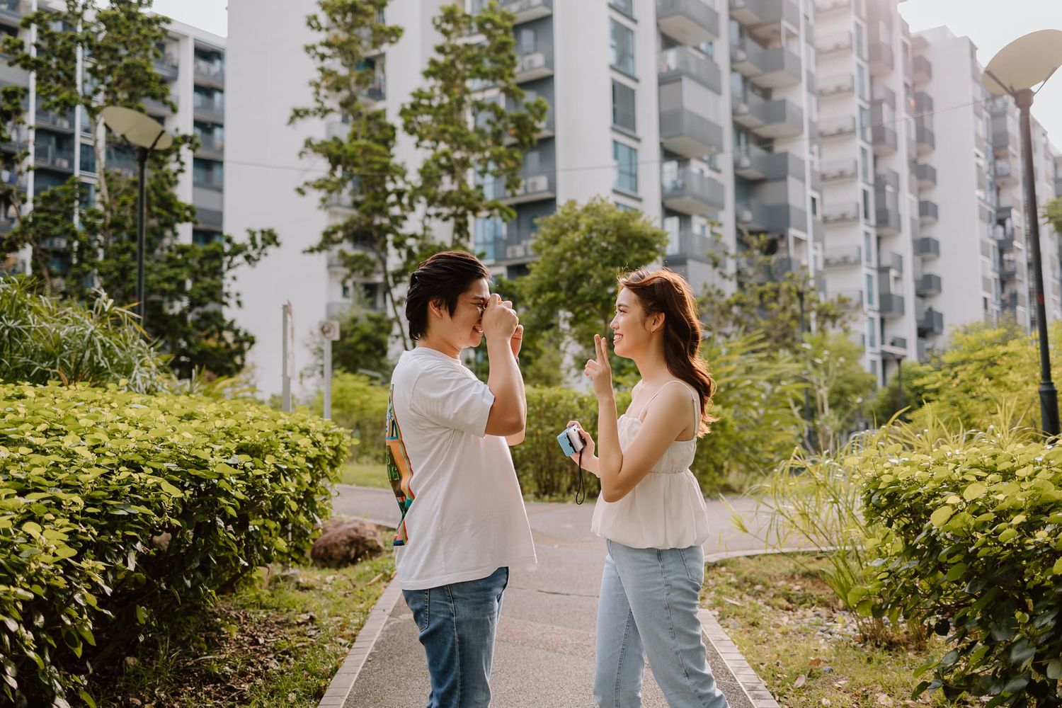 Playful couple in white tops and jeans high-fiving each other in front of apartment buildings.