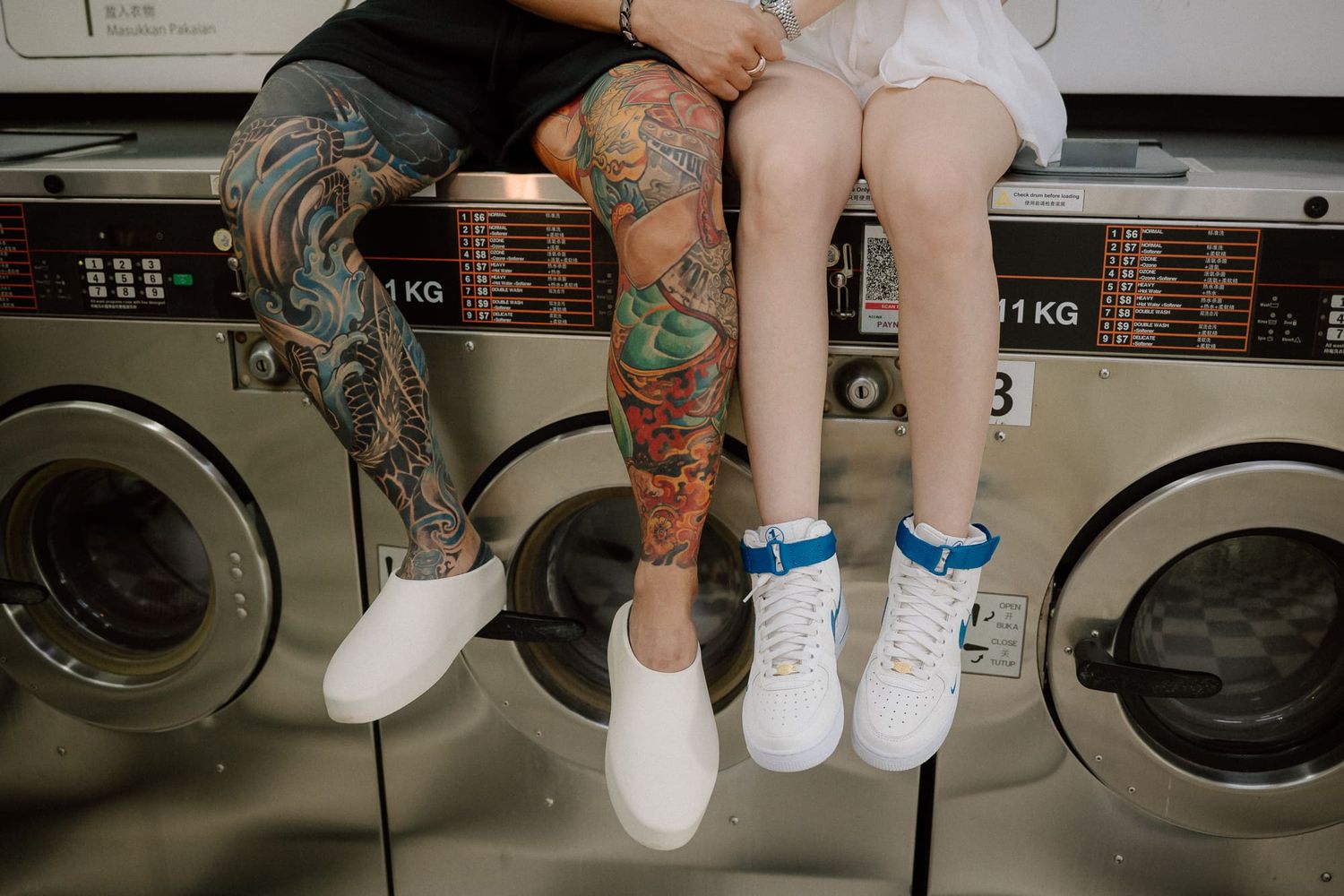 Two pairs of legs with colorful tattoos and white sneakers dangling in front of industrial washing machines at a laundromat.